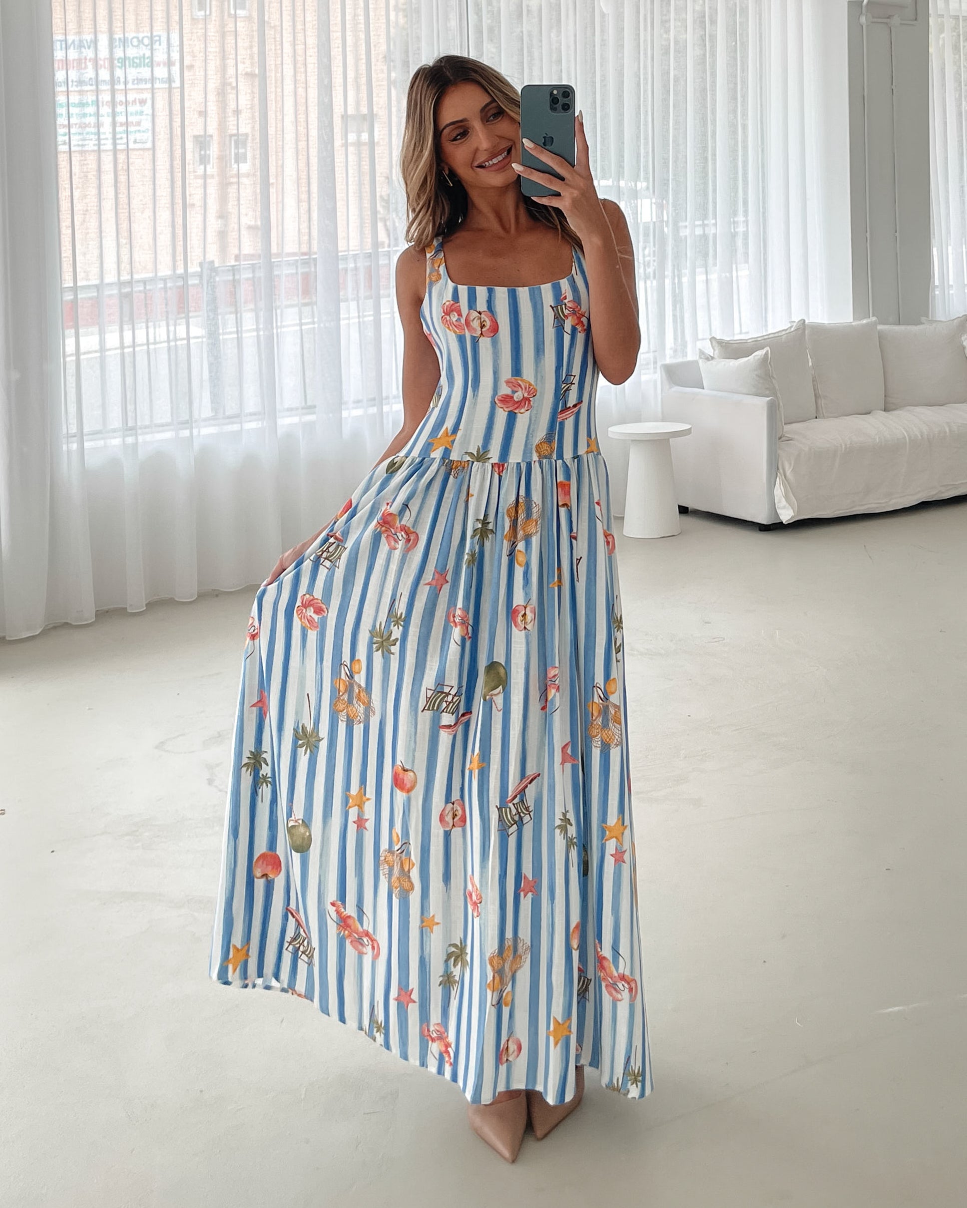 A model wearing a white and blue base multicolored print maxi dress with a square neckline and wide straps.