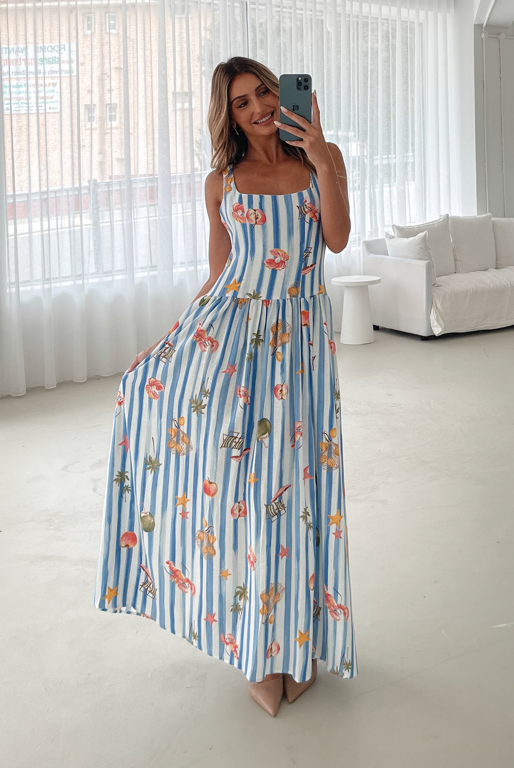 A model wearing a white and blue base multicolored print maxi dress with a square neckline and wide straps.