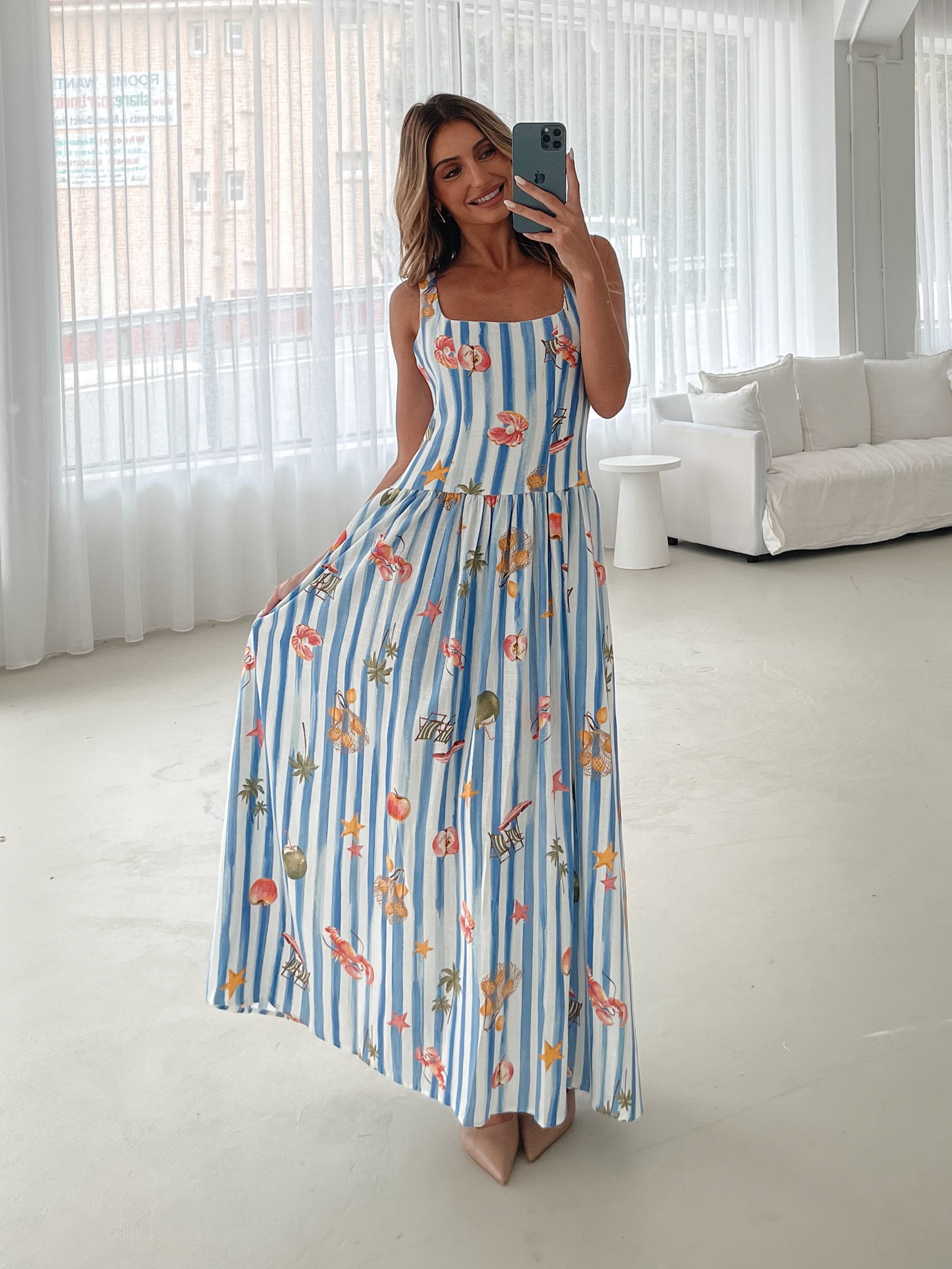 A model wearing a white and blue base multicolored print maxi dress with a square neckline and wide straps.