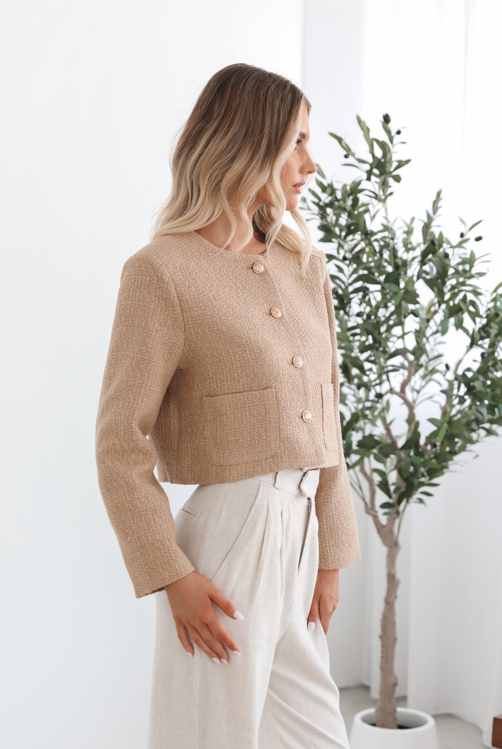 A woman wearing a beige Jacket with long sleeves, round neckline, and center-front buttons. The jacket has a crop length and she is paired with light-colored trousers.