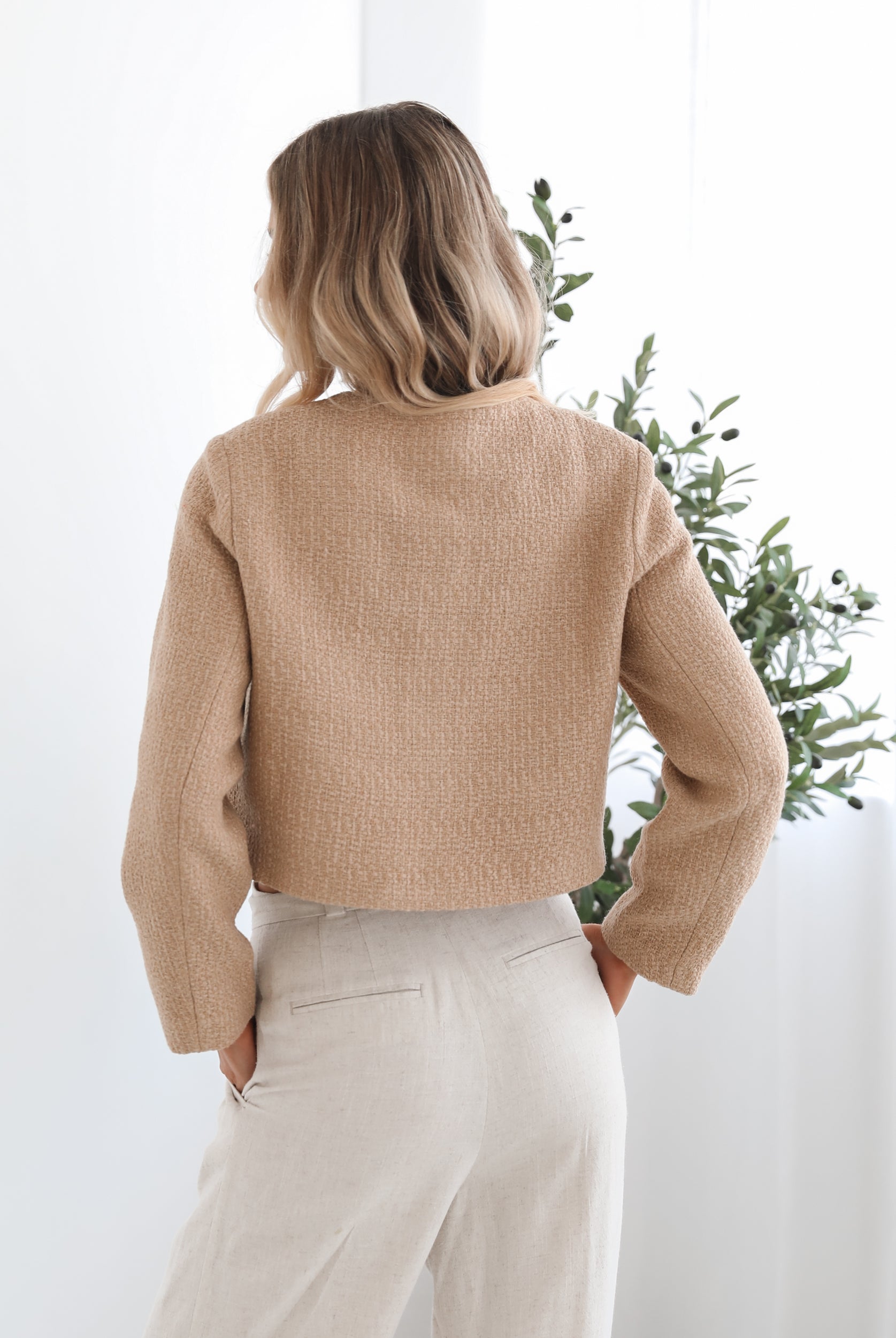 A woman wearing a beige Jacket with long sleeves, round neckline, and center-front buttons. The jacket has a crop length and she is paired with light-colored trousers.