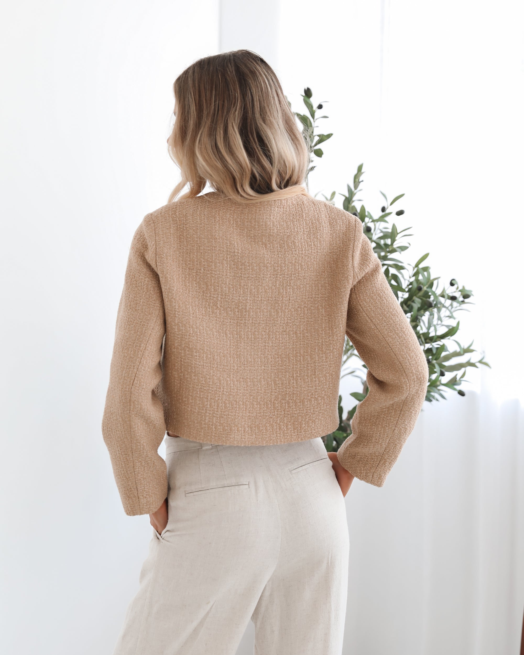 A woman wearing a beige Jacket with long sleeves, round neckline, and center-front buttons. The jacket has a crop length and she is paired with light-colored trousers.