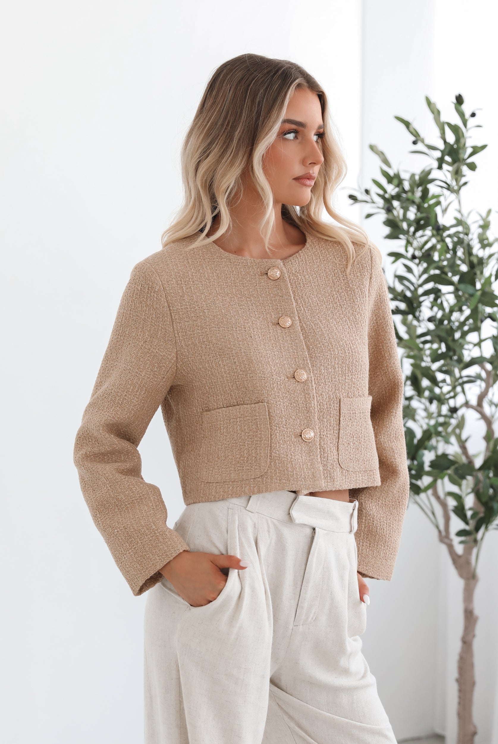 A woman wearing a beige Jacket with long sleeves, round neckline, and center-front buttons. The jacket has a crop length and she is paired with light-colored trousers.