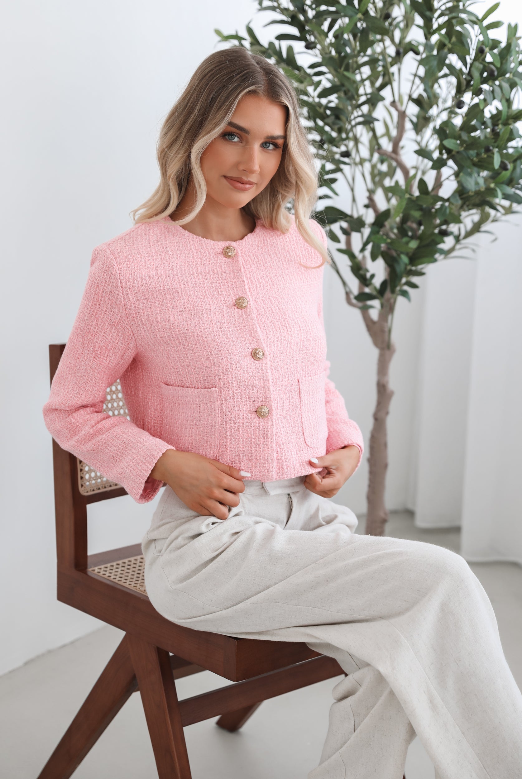 A woman wearing a pink Valdivia Jacket with long sleeves, round neckline, and center-front buttons. The jacket has a crop length and she is paired with light-colored trousers.