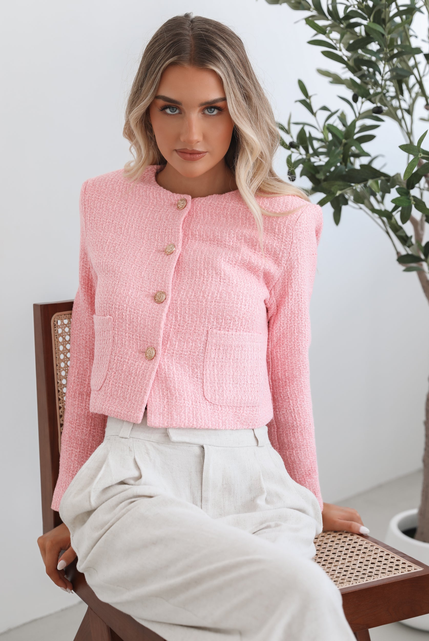 A woman wearing a pink Valdivia Jacket with long sleeves, round neckline, and center-front buttons. The jacket has a crop length and she is paired with light-colored trousers.