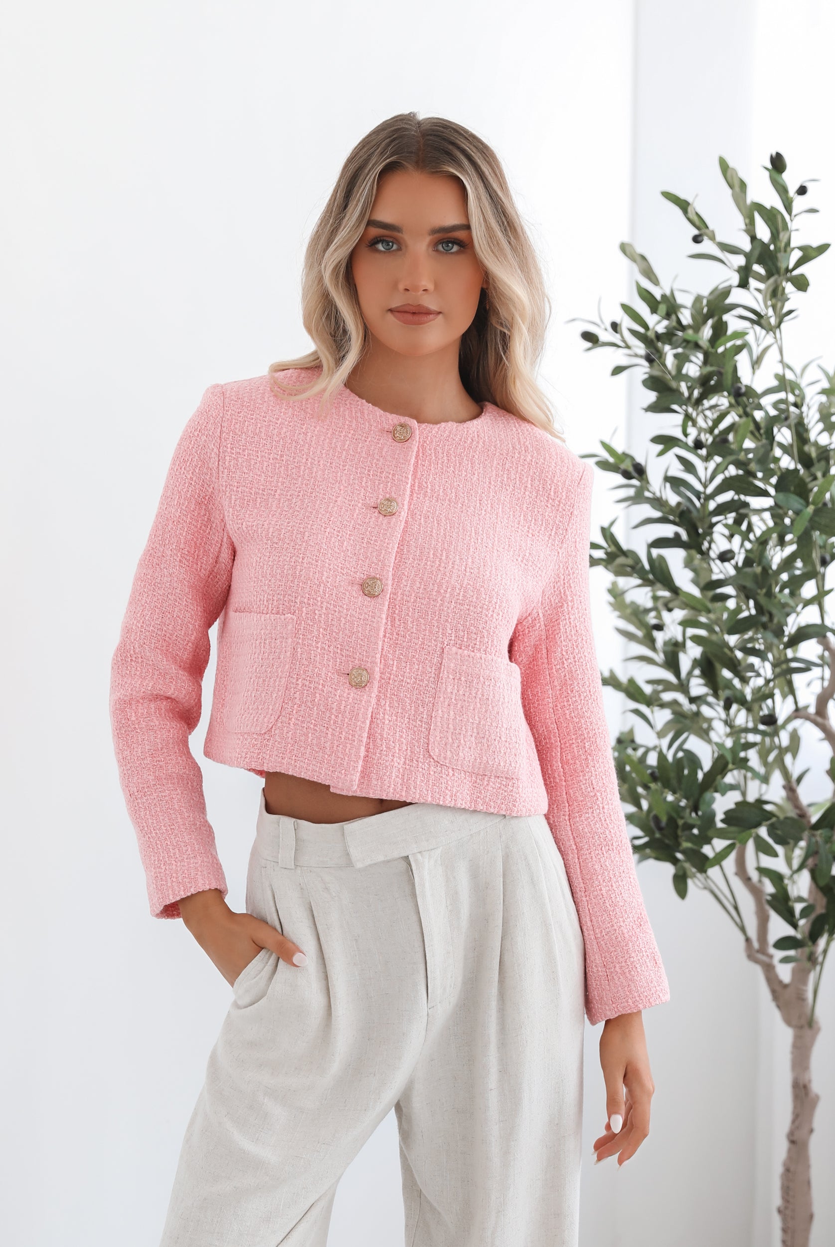 A woman wearing a pink Valdivia Jacket with long sleeves, round neckline, and center-front buttons. The jacket has a crop length and she is paired with light-colored trousers.