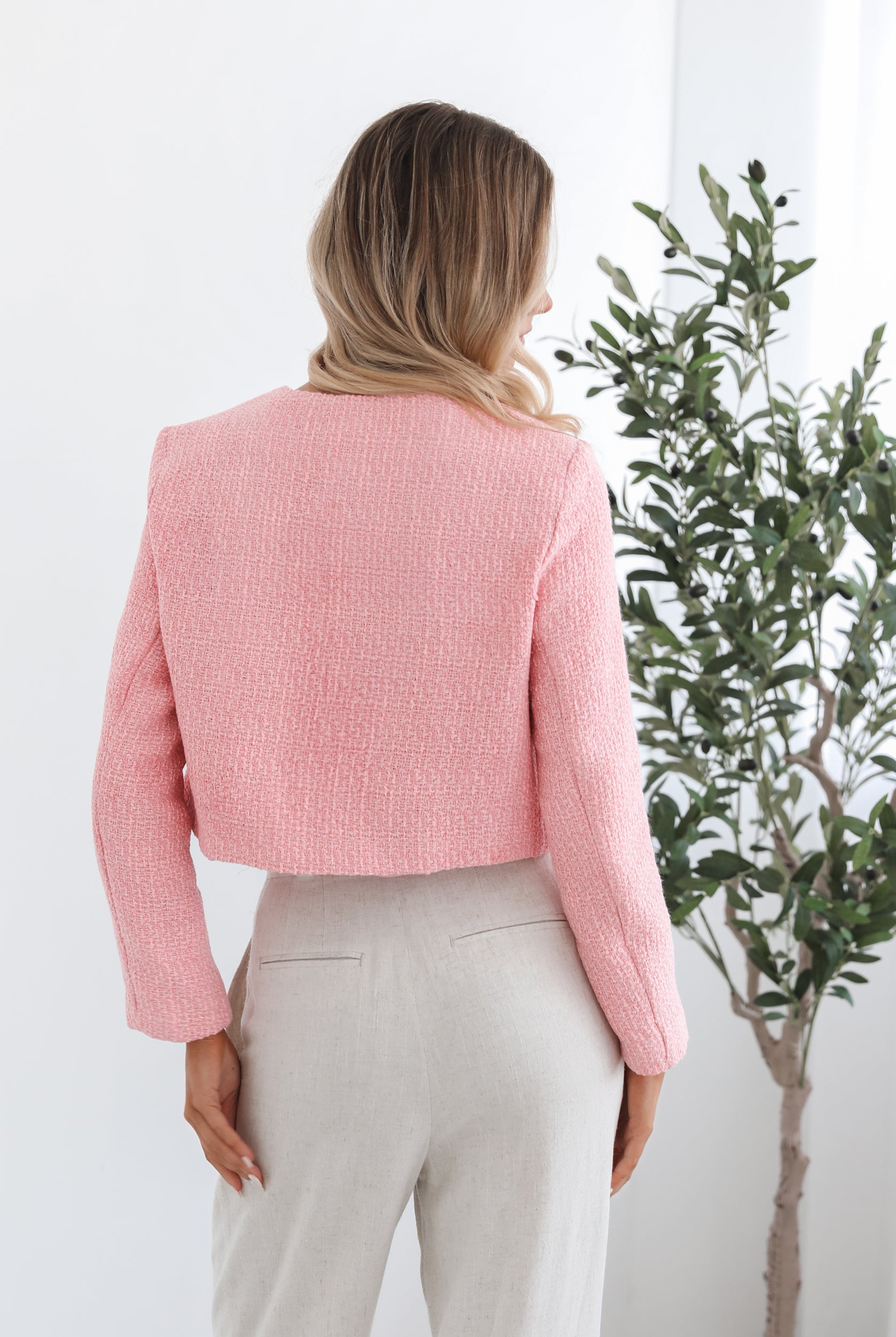 A woman wearing a pink Valdivia Jacket with long sleeves, round neckline, and center-front buttons. The jacket has a crop length and she is paired with light-colored trousers.