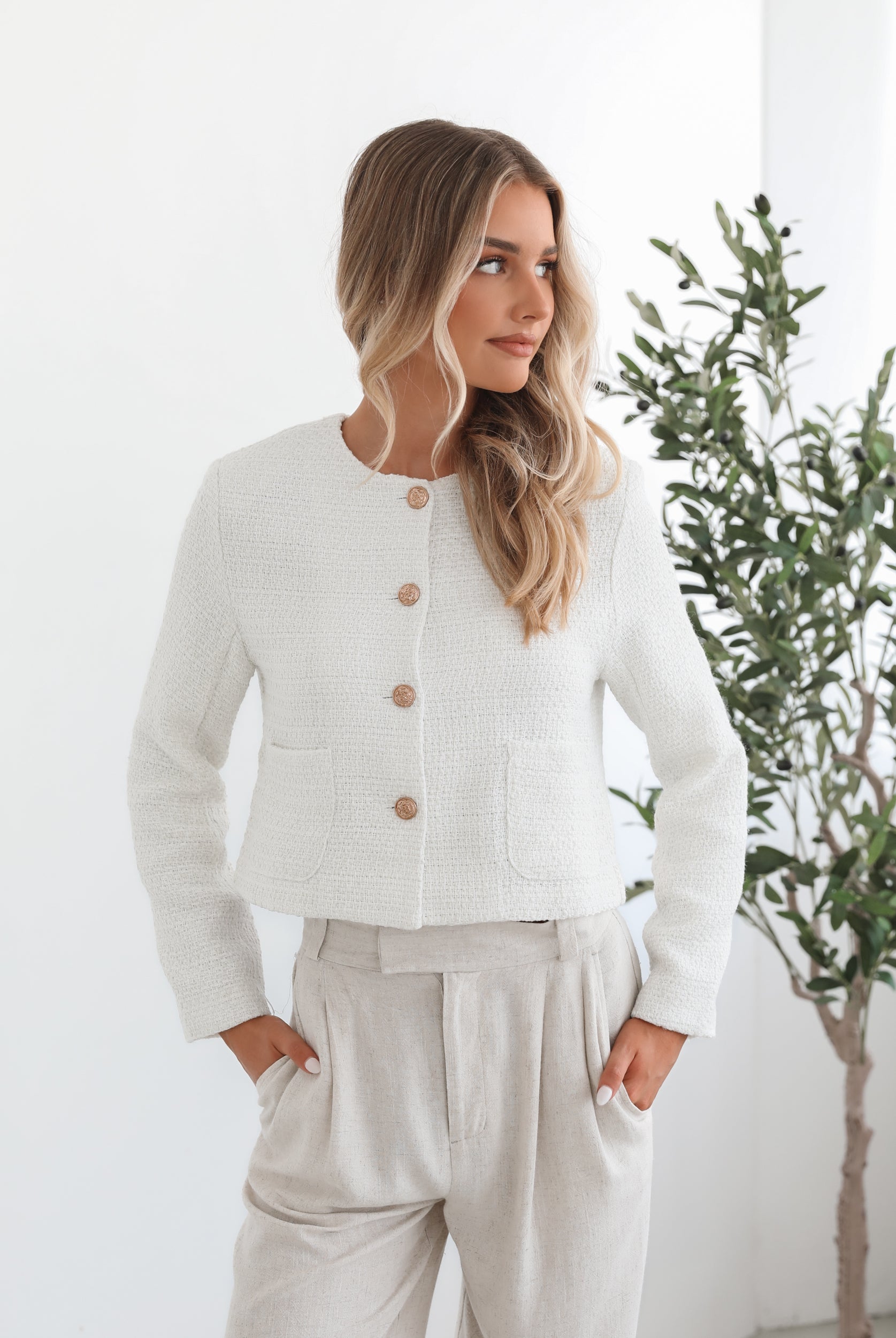 A woman sitting in a chair wearing a white Valdivia Jacket with long sleeves, round neckline, and center-front buttons. The jacket has a crop length and she is paired with a light-colored pants.