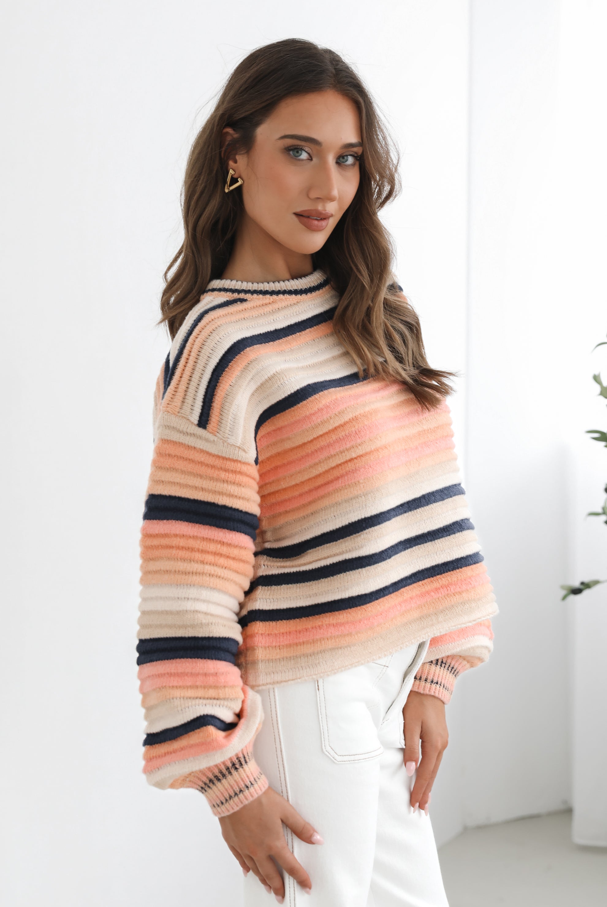 A woman sitting in a chair wearing a striped peach, pink, blue, and black knit top with a round neckline and long sleeves, paired with white pants.
