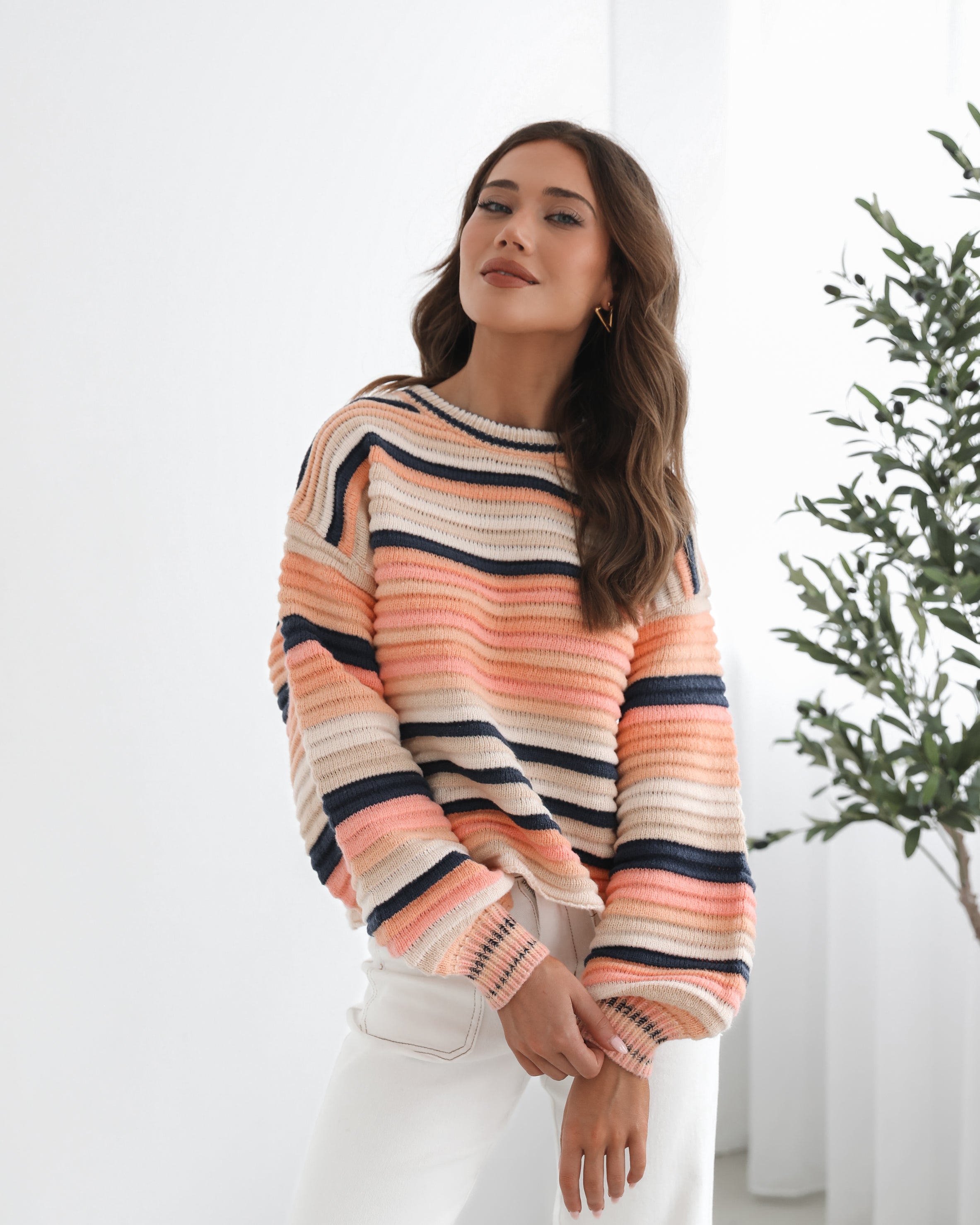 A woman sitting in a chair wearing a striped peach, pink, blue, and black knit top with a round neckline and long sleeves, paired with white pants.