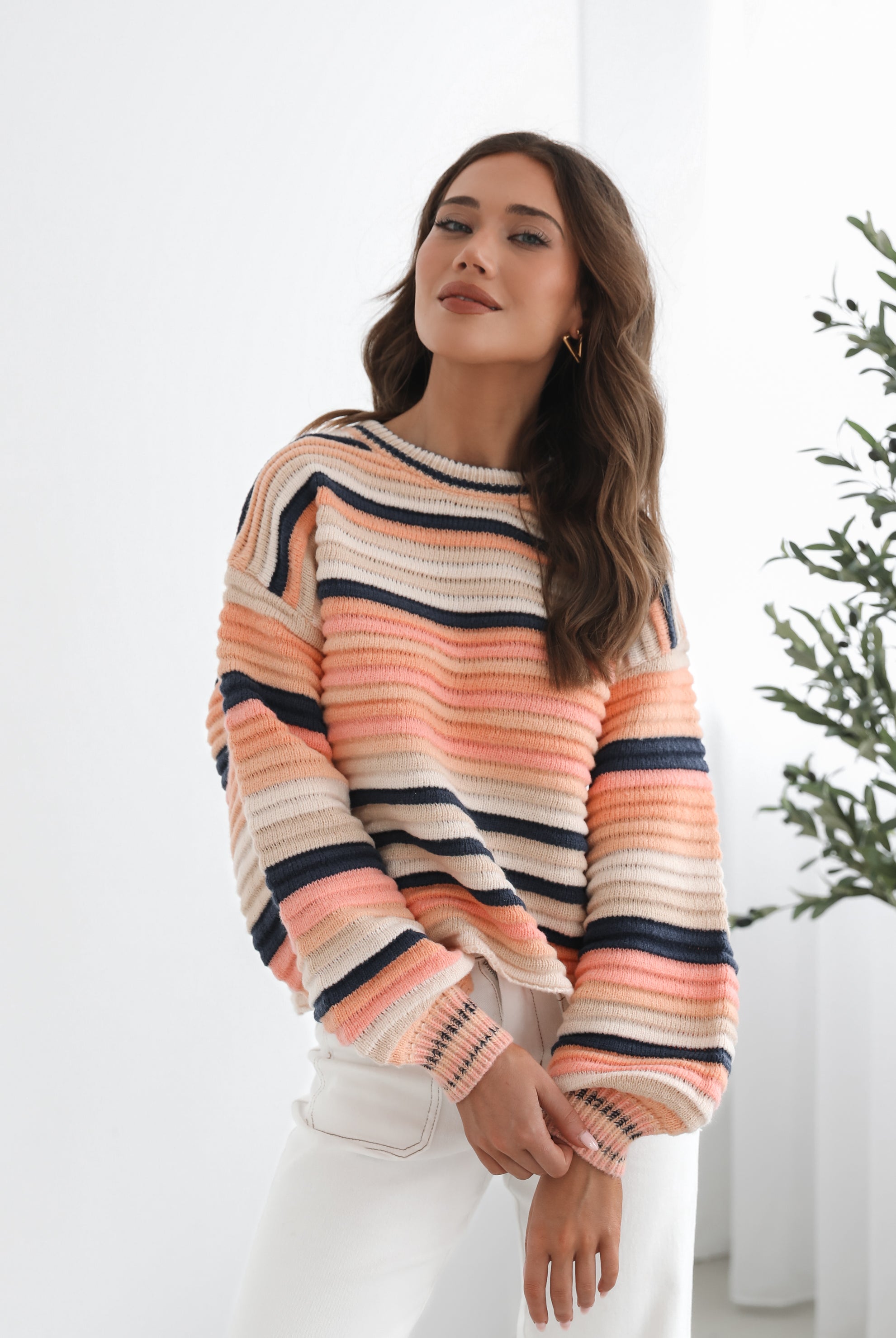 A woman sitting in a chair wearing a striped peach, pink, blue, and black knit top with a round neckline and long sleeves, paired with white pants.