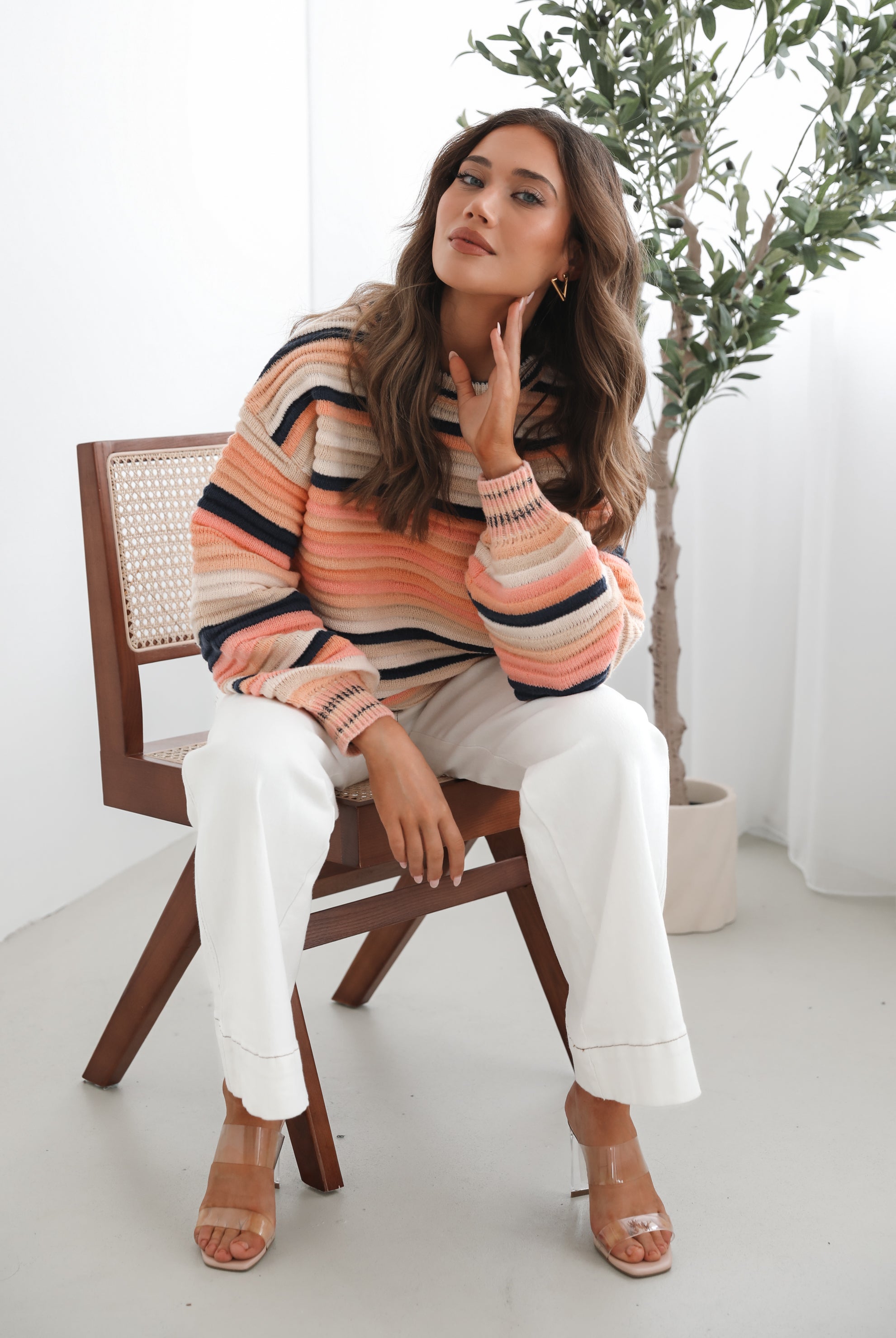 A woman sitting in a chair wearing a striped peach, pink, blue, and black knit top with a round neckline and long sleeves, paired with white pants.