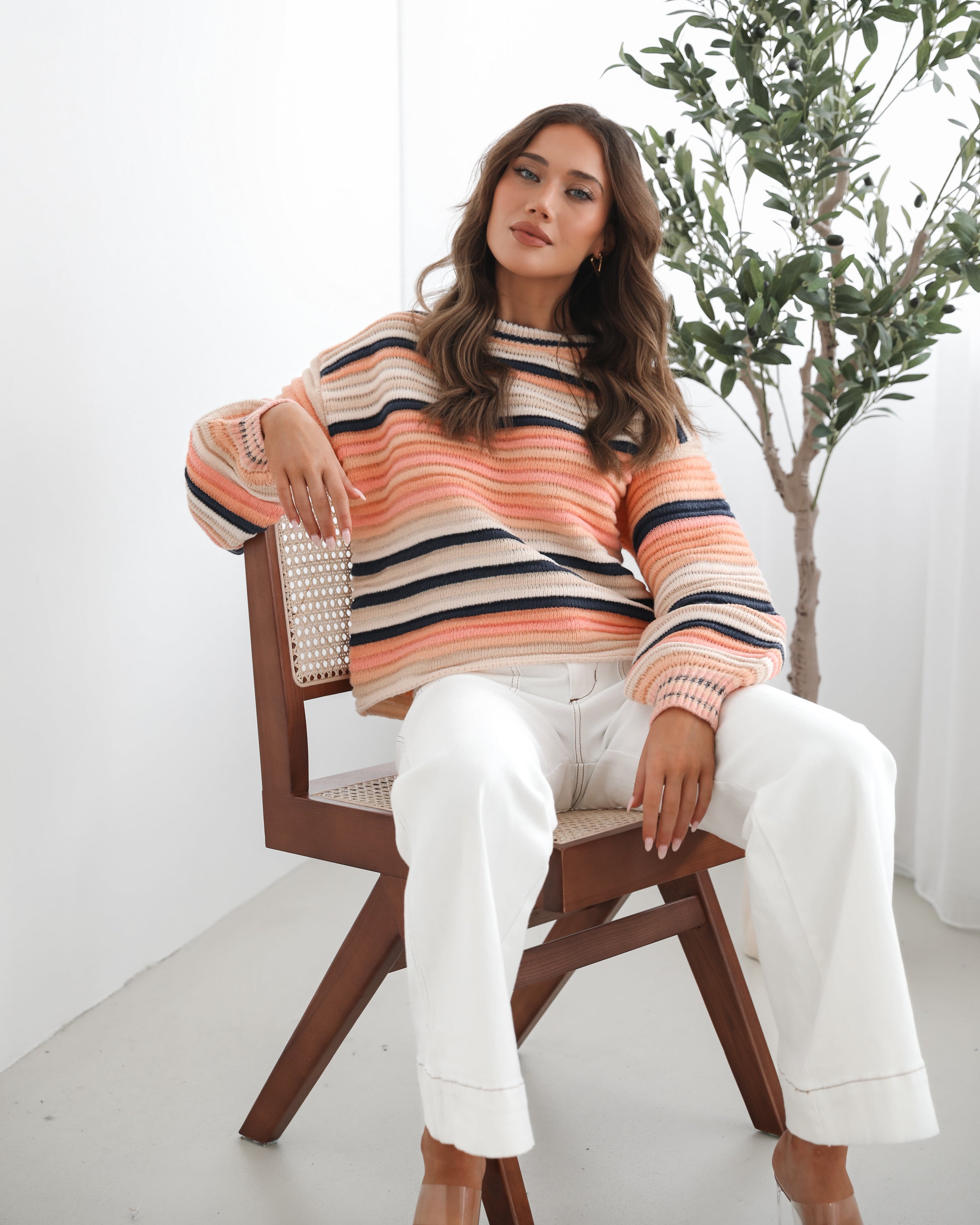 A woman sitting in a chair wearing a striped peach, pink, blue, and black knit top with a round neckline and long sleeves, paired with white pants.