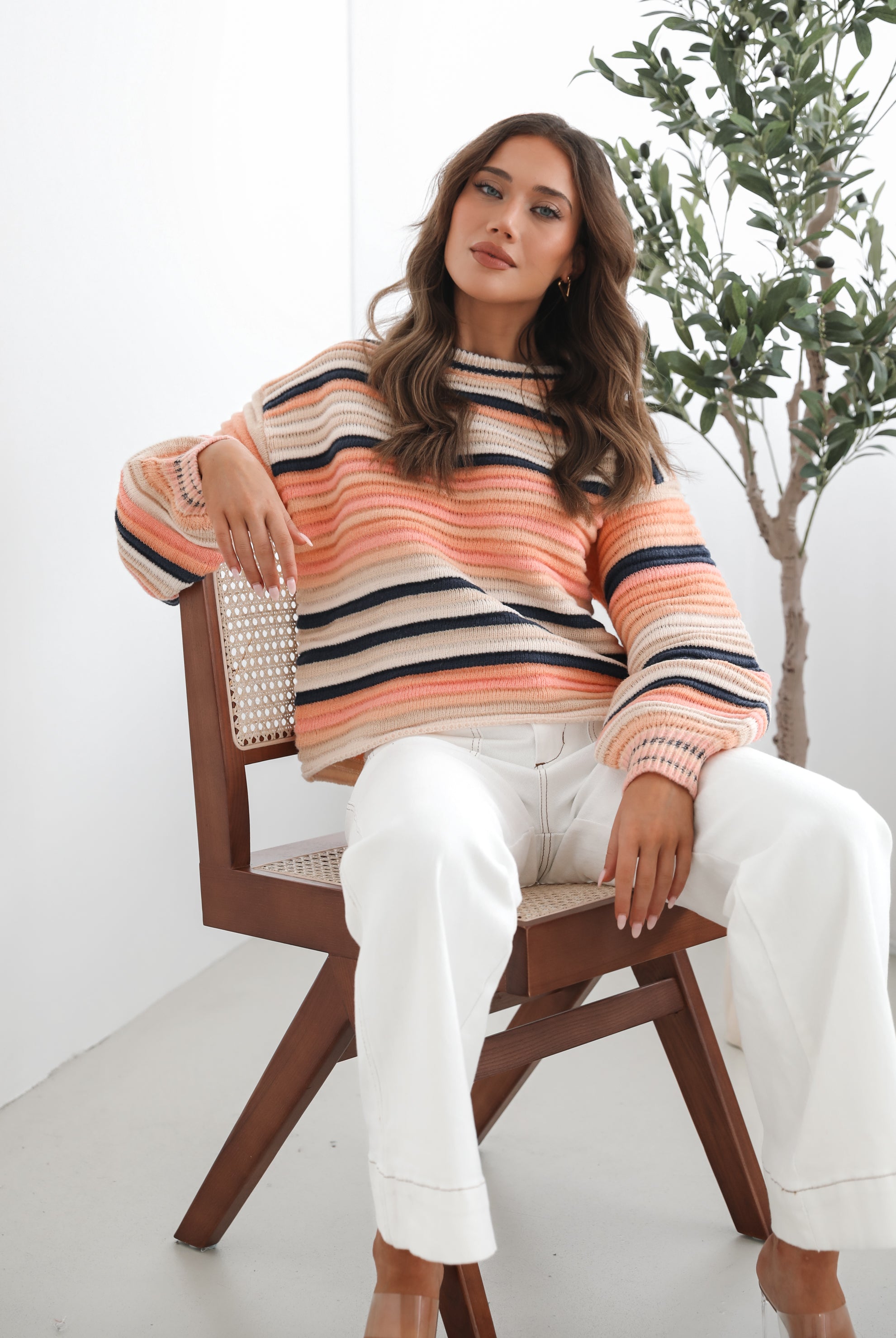 A woman sitting in a chair wearing a striped peach, pink, blue, and black knit top with a round neckline and long sleeves, paired with white pants.