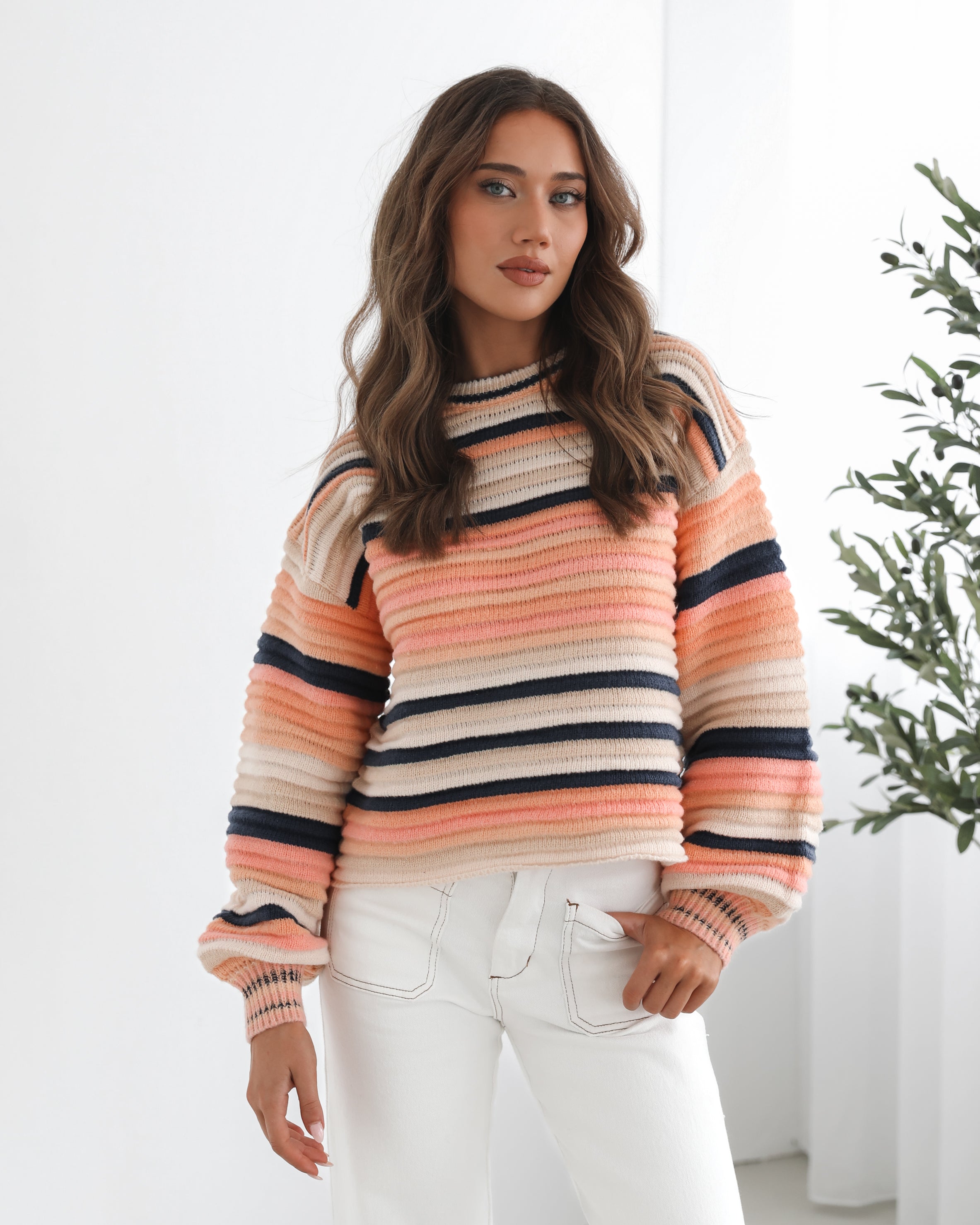 A woman sitting in a chair wearing a striped peach, pink, blue, and black knit top with a round neckline and long sleeves, paired with white pants.