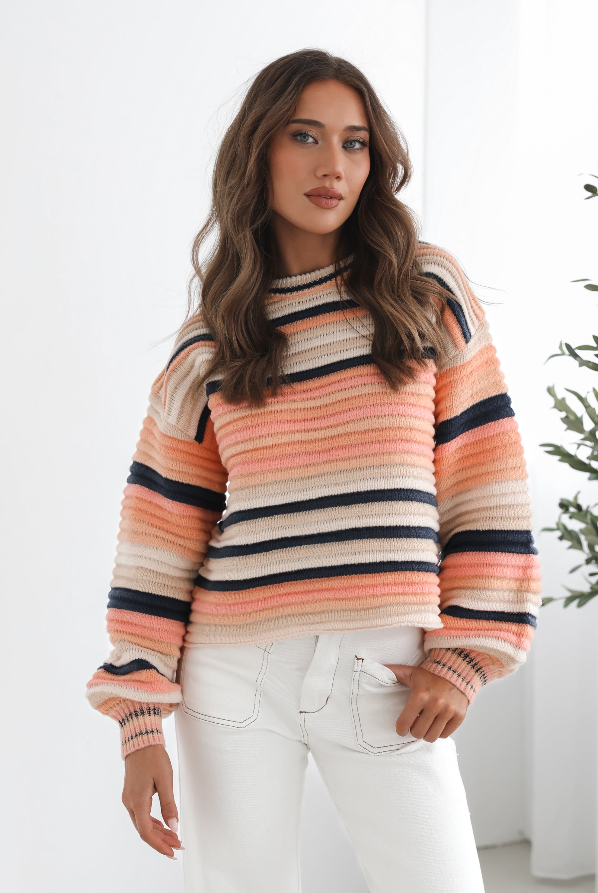 A woman sitting in a chair wearing a striped peach, pink, blue, and black knit top with a round neckline and long sleeves, paired with white pants.