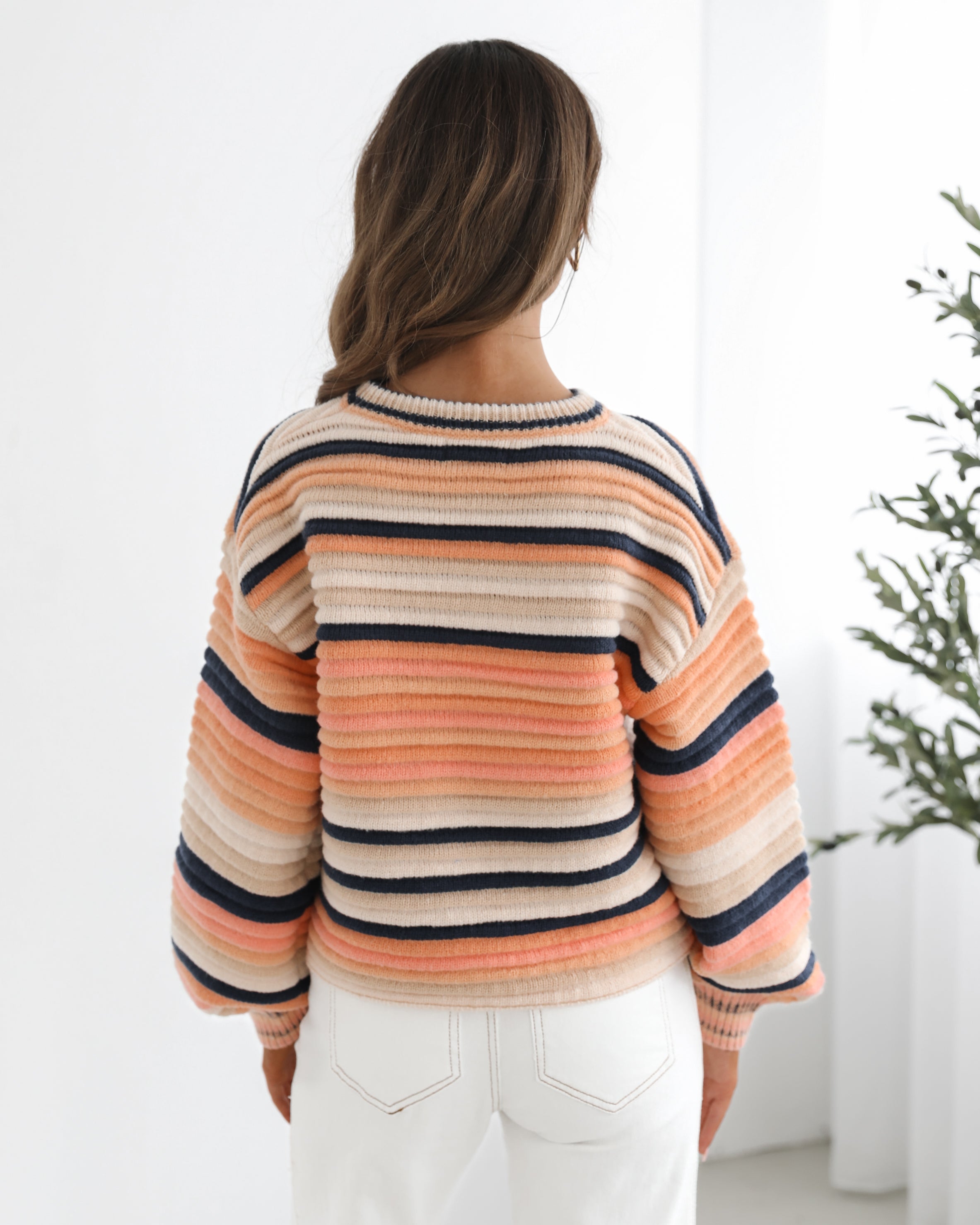 A woman sitting in a chair wearing a striped peach, pink, blue, and black knit top with a round neckline and long sleeves, paired with white pants.