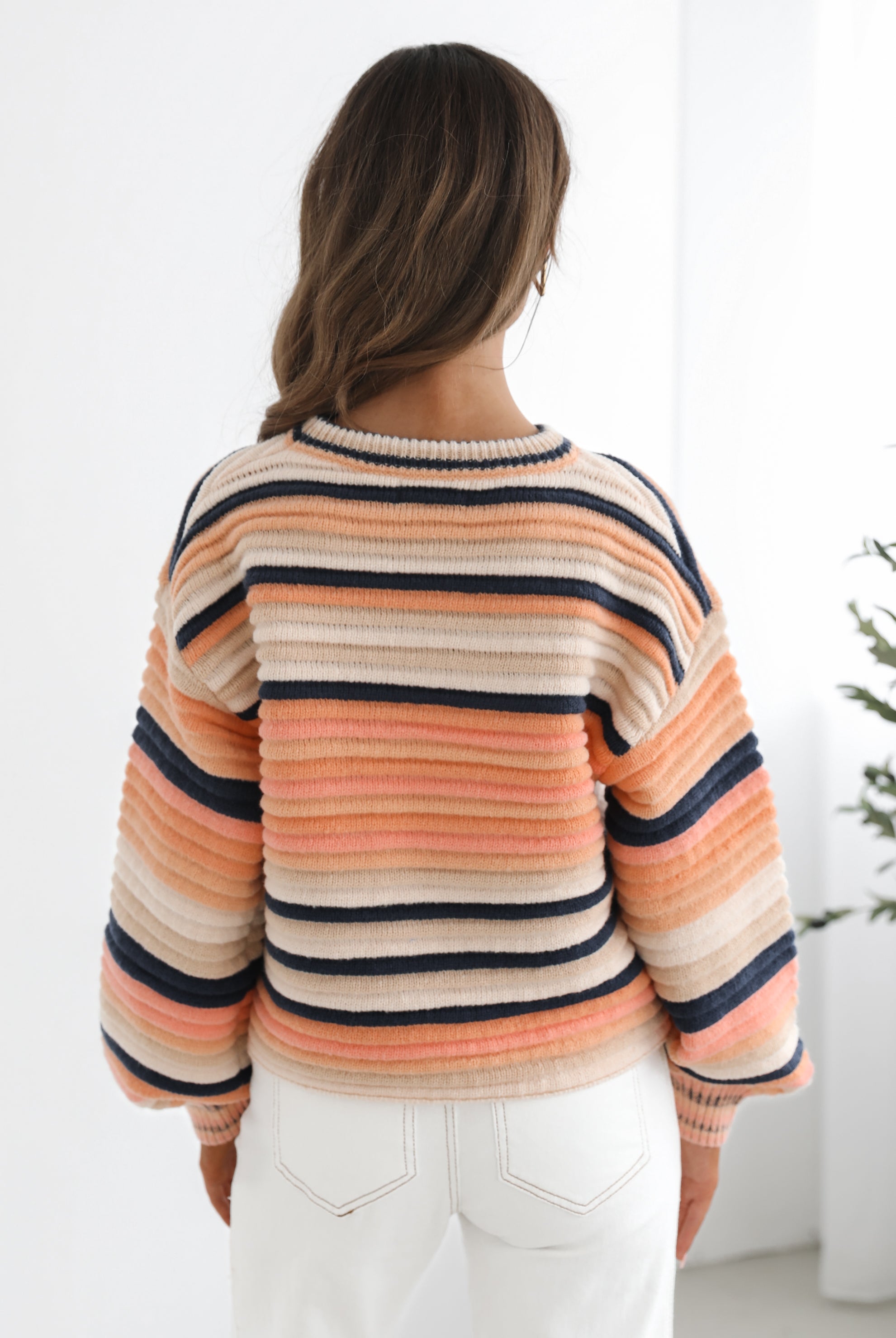 A woman sitting in a chair wearing a striped peach, pink, blue, and black knit top with a round neckline and long sleeves, paired with white pants.