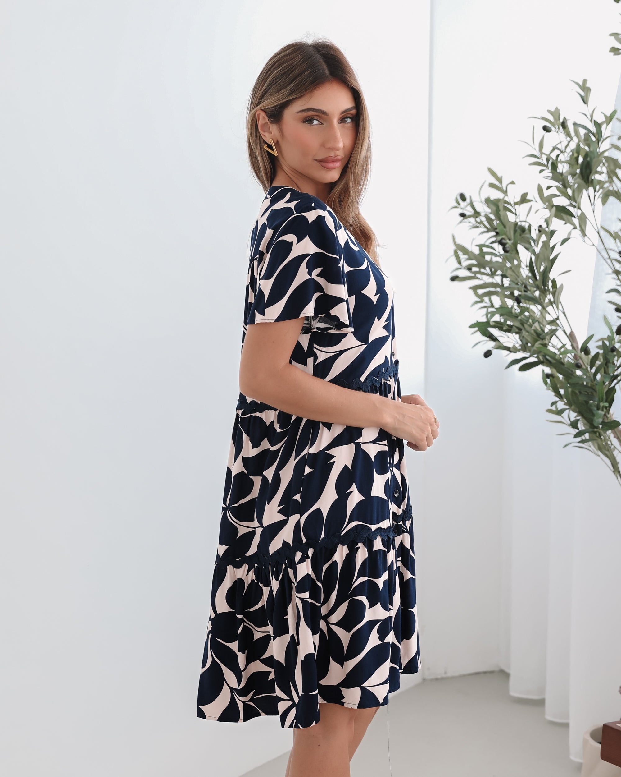 A woman standing in front of a plant, wearing a beige dress with navy-tone leaf prints, short flutter sleeves, and a v-neckline.