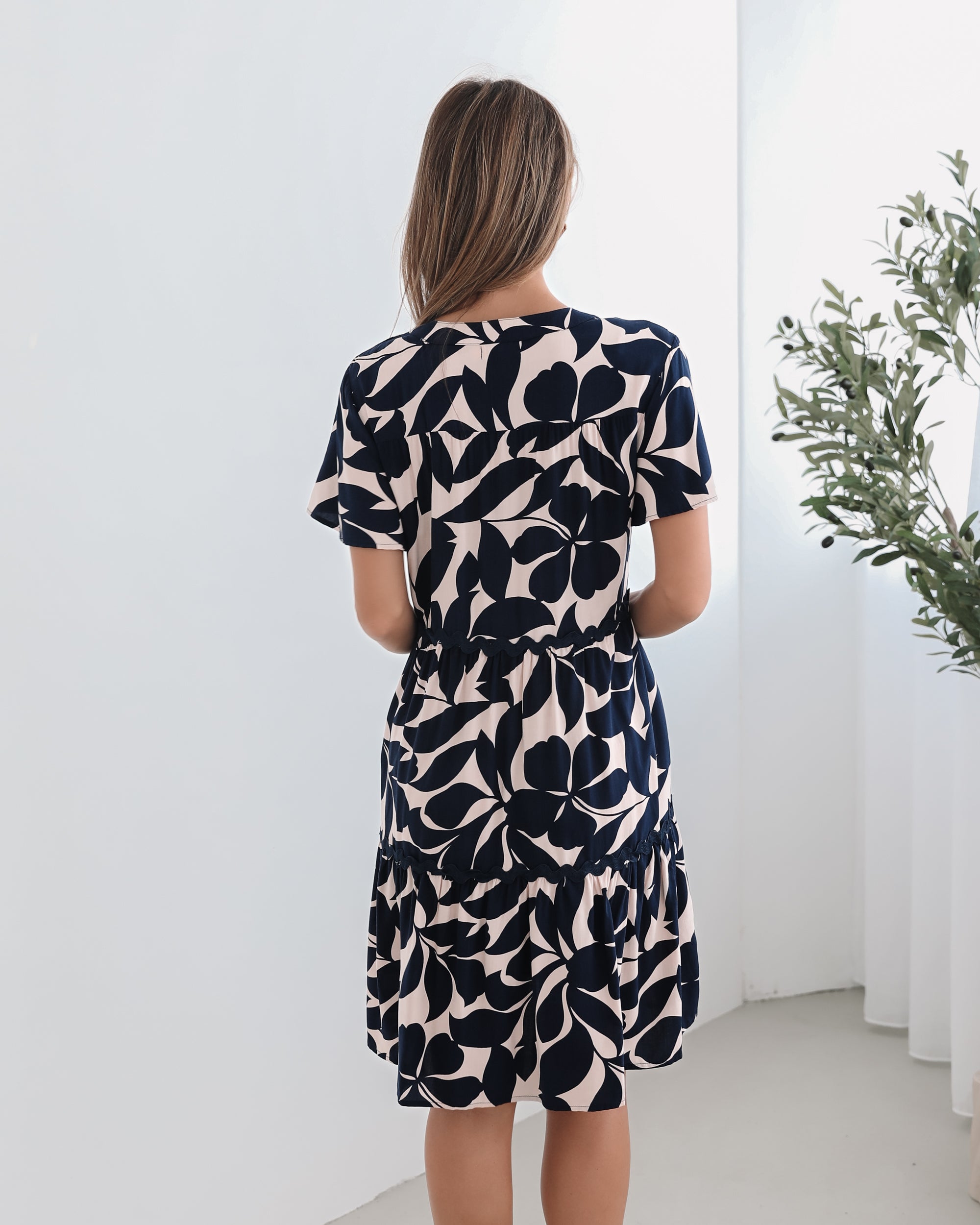 A woman standing in front of a plant, wearing a beige dress with navy-tone leaf prints, short flutter sleeves, and a v-neckline.
