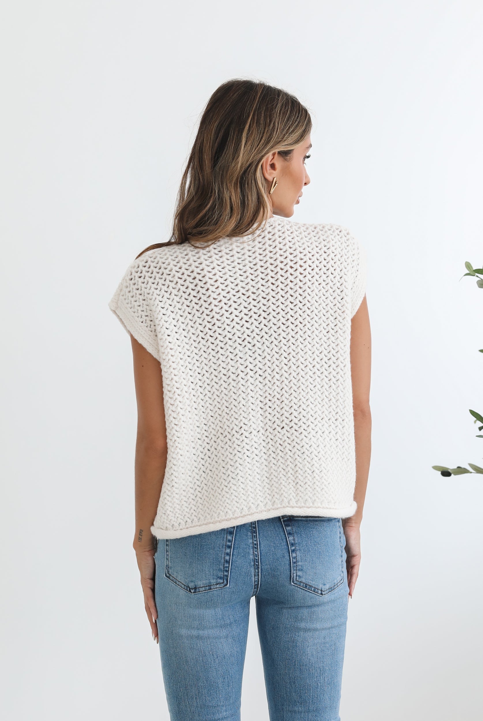 A woman wearing a beige knit top with short sleeves, a round neckline, and center-front buttons. She is paired with blue jeans and is standing in a room with a plant in the background.