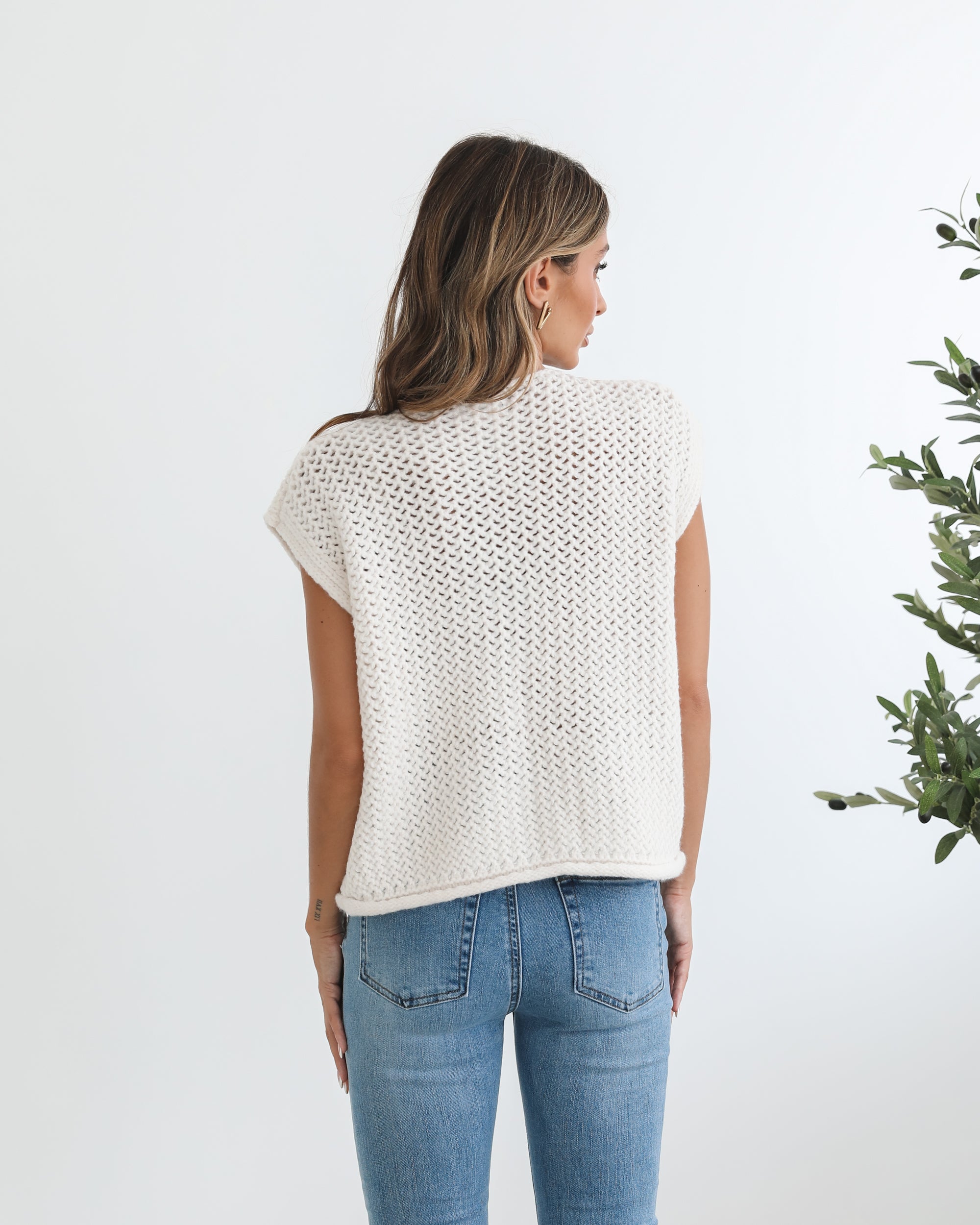 A woman wearing a beige knit top with short sleeves, a round neckline, and center-front buttons. She is paired with blue jeans and is standing in a room with a plant in the background.