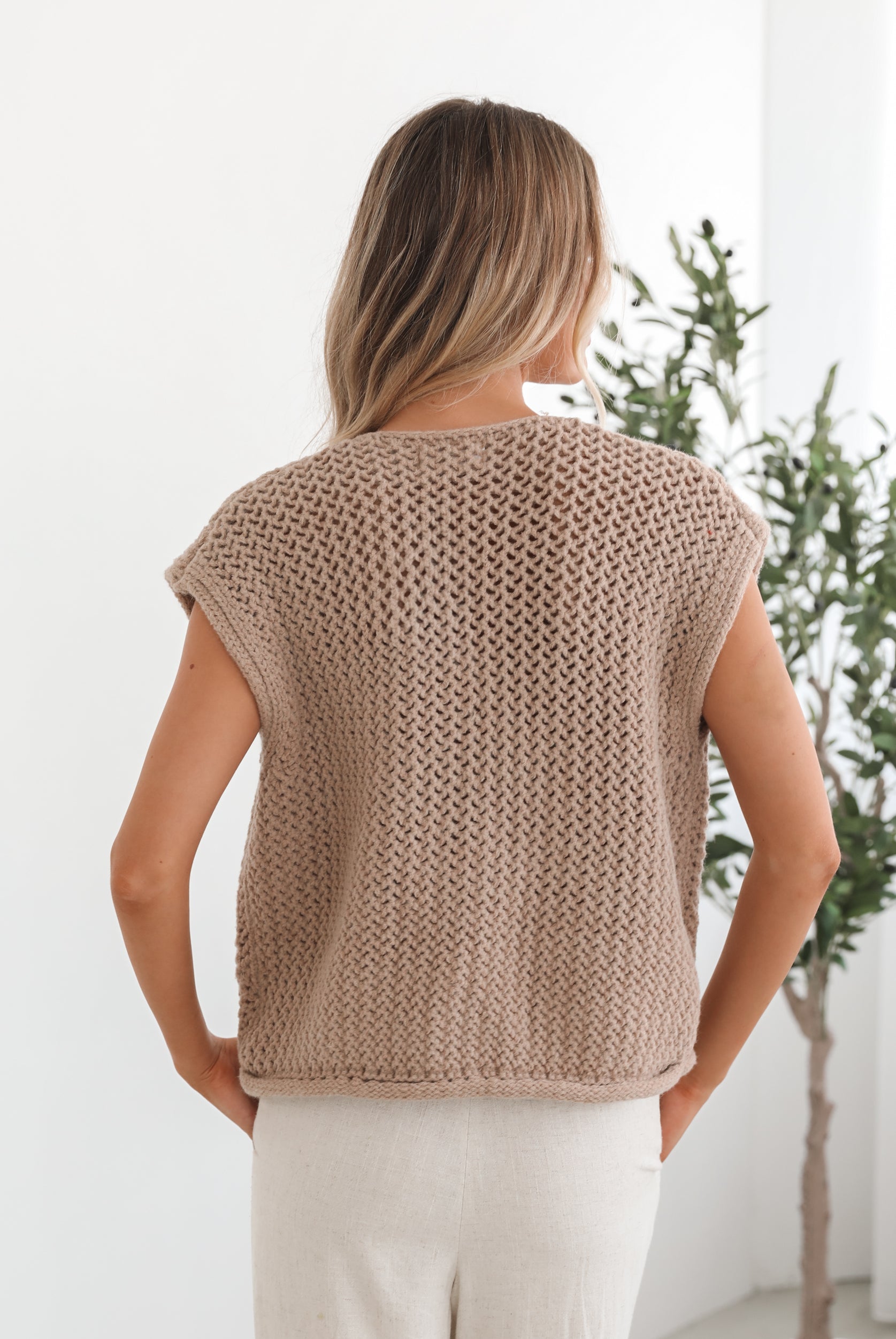A woman wearing a beige knit top with round neckline, centre-front buttons, and a relaxed fit. The top is sleeveless and she is pairing it with light-colored pants.