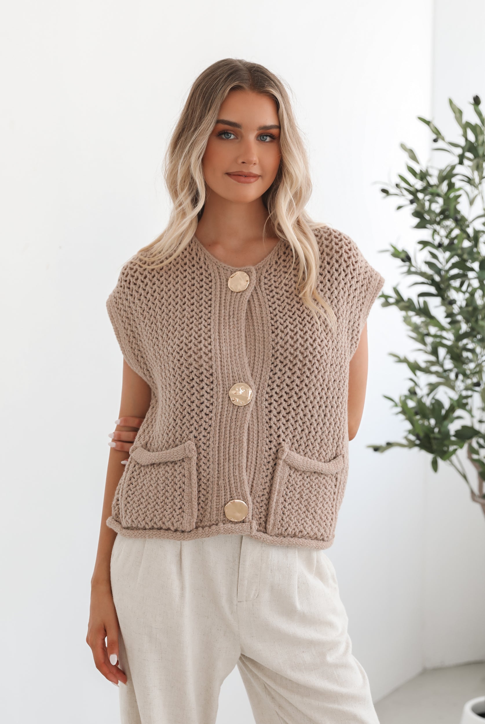 A woman wearing a beige knit top with round neckline, centre-front buttons, and a relaxed fit. The top is sleeveless and she is pairing it with light-colored pants.