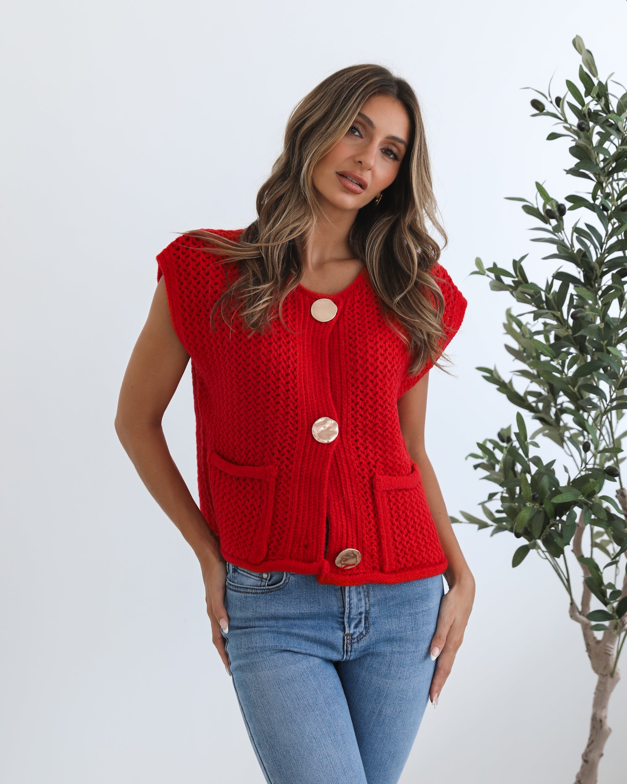 A woman wearing a red knit top with round neckline and center-front buttons, paired with blue jeans.