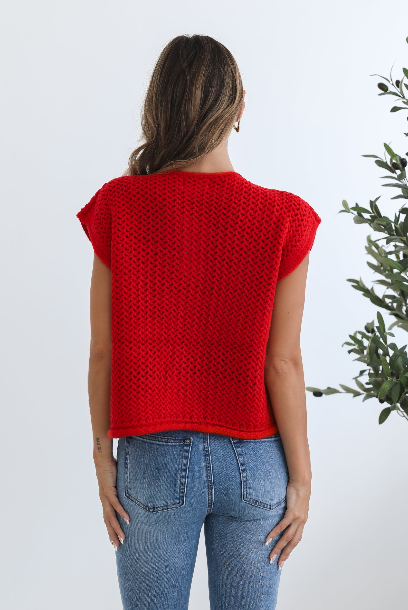 A woman wearing a red knit top with round neckline and center-front buttons, paired with blue jeans.