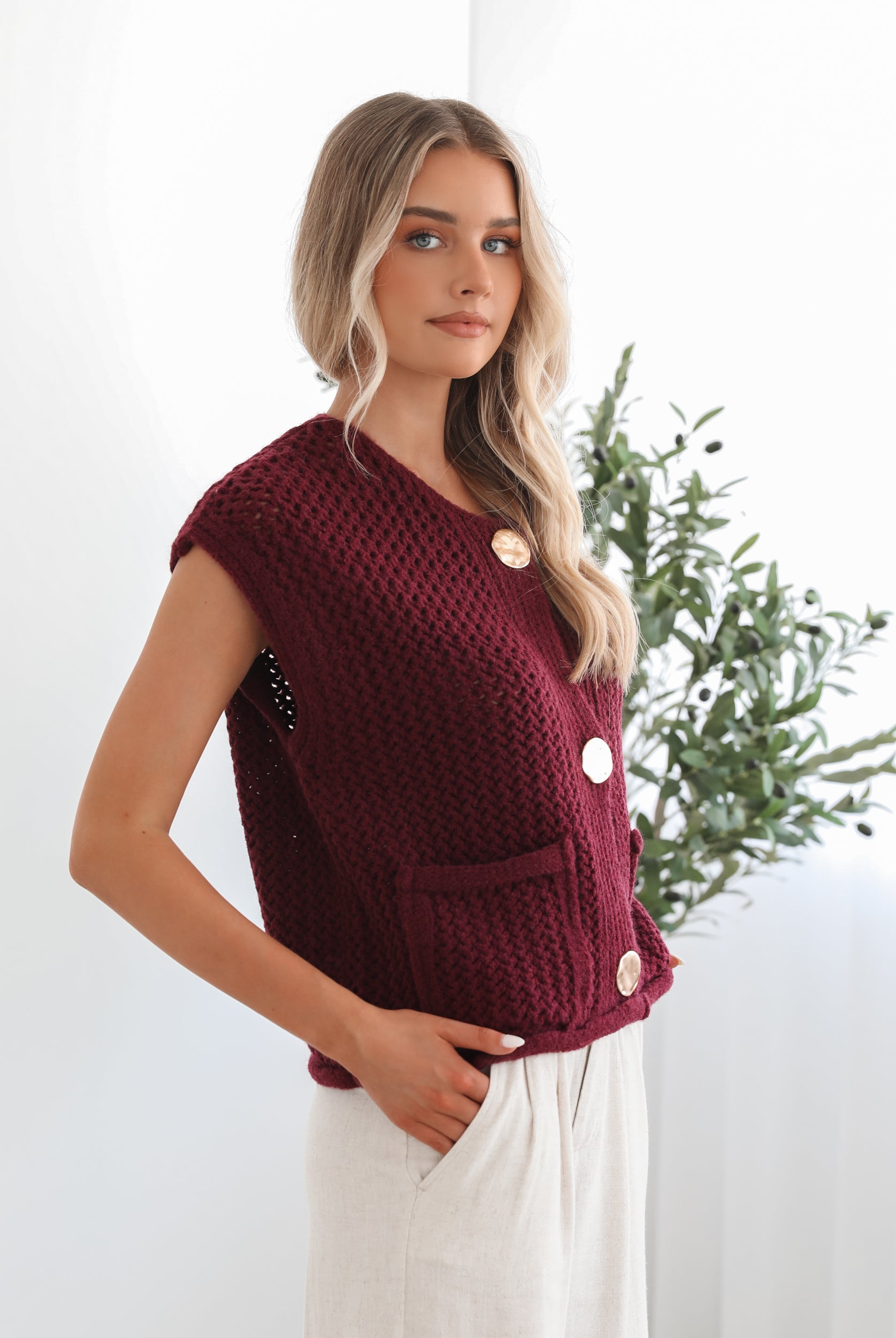 A woman wearing a sleeveless wine-colored knit top with round neckline and center-front buttons, paired with light-colored pants.