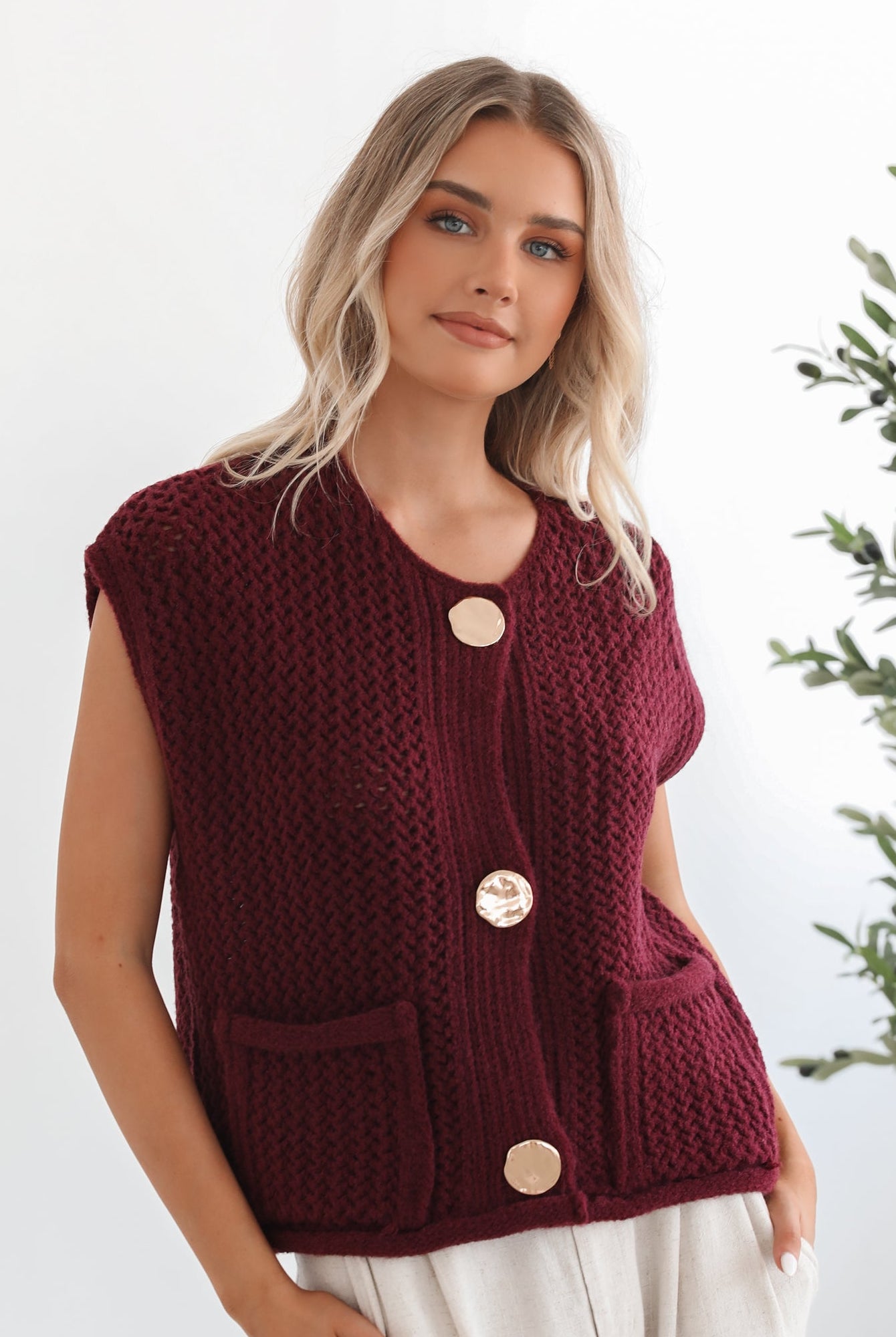A woman wearing a sleeveless wine-colored knit top with round neckline and center-front buttons, paired with light-colored pants.