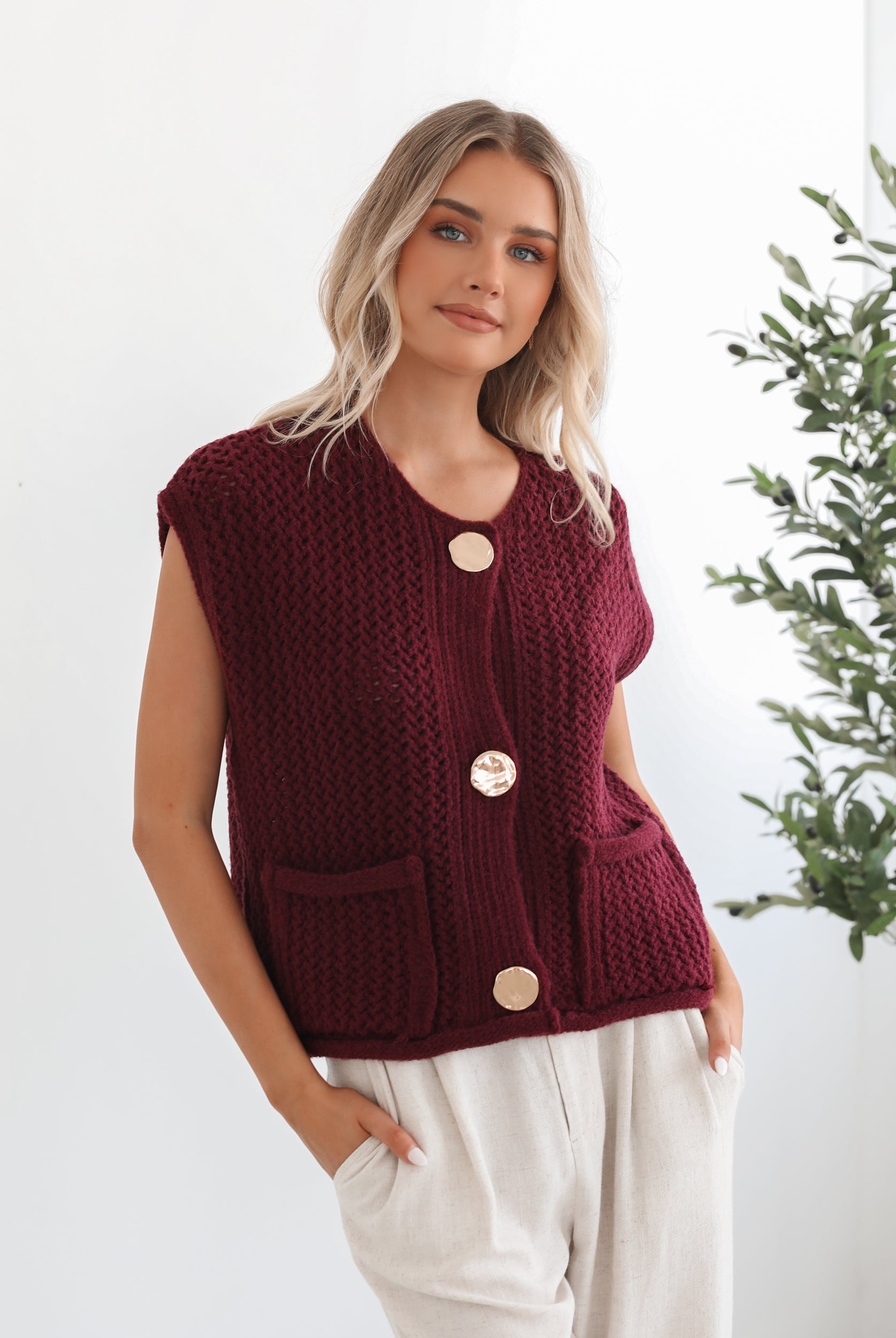A woman wearing a sleeveless wine-colored knit top with round neckline and center-front buttons, paired with light-colored pants.