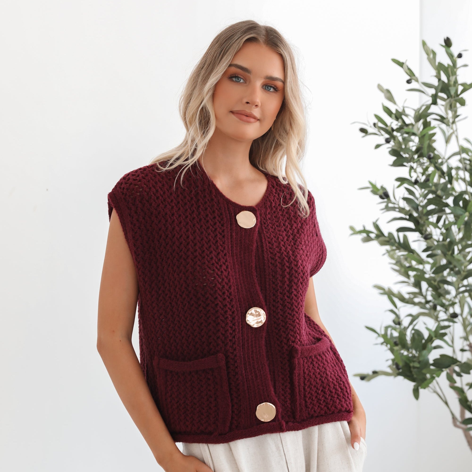 A woman wearing a sleeveless wine-colored knit top with round neckline and center-front buttons, paired with light-colored pants.