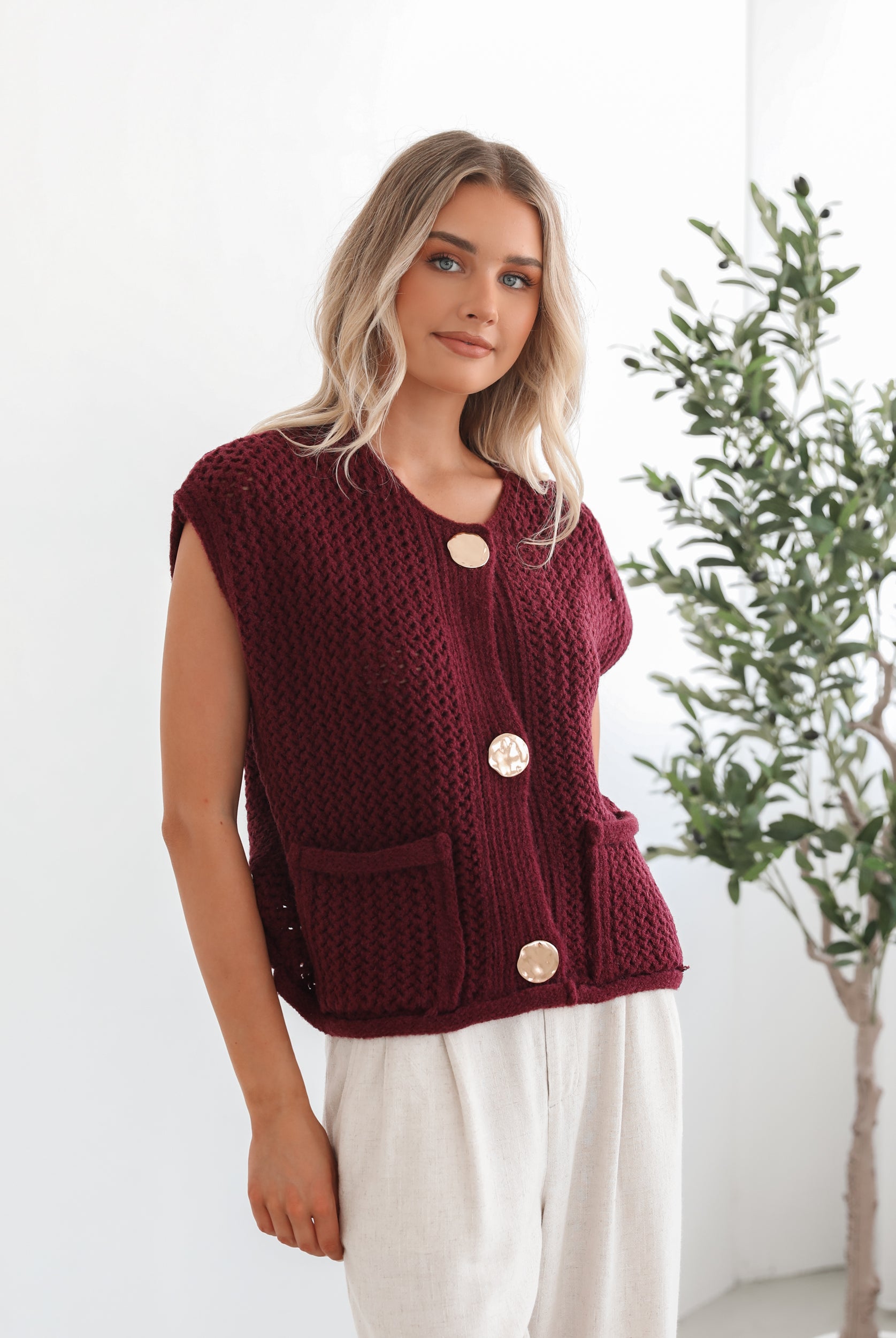 A woman wearing a sleeveless wine-colored knit top with round neckline and center-front buttons, paired with light-colored pants.