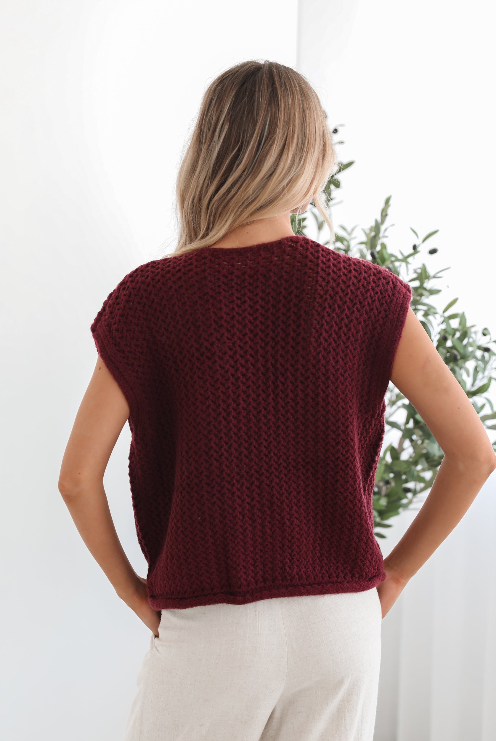 A woman wearing a sleeveless wine-colored knit top with round neckline and center-front buttons, paired with light-colored pants.