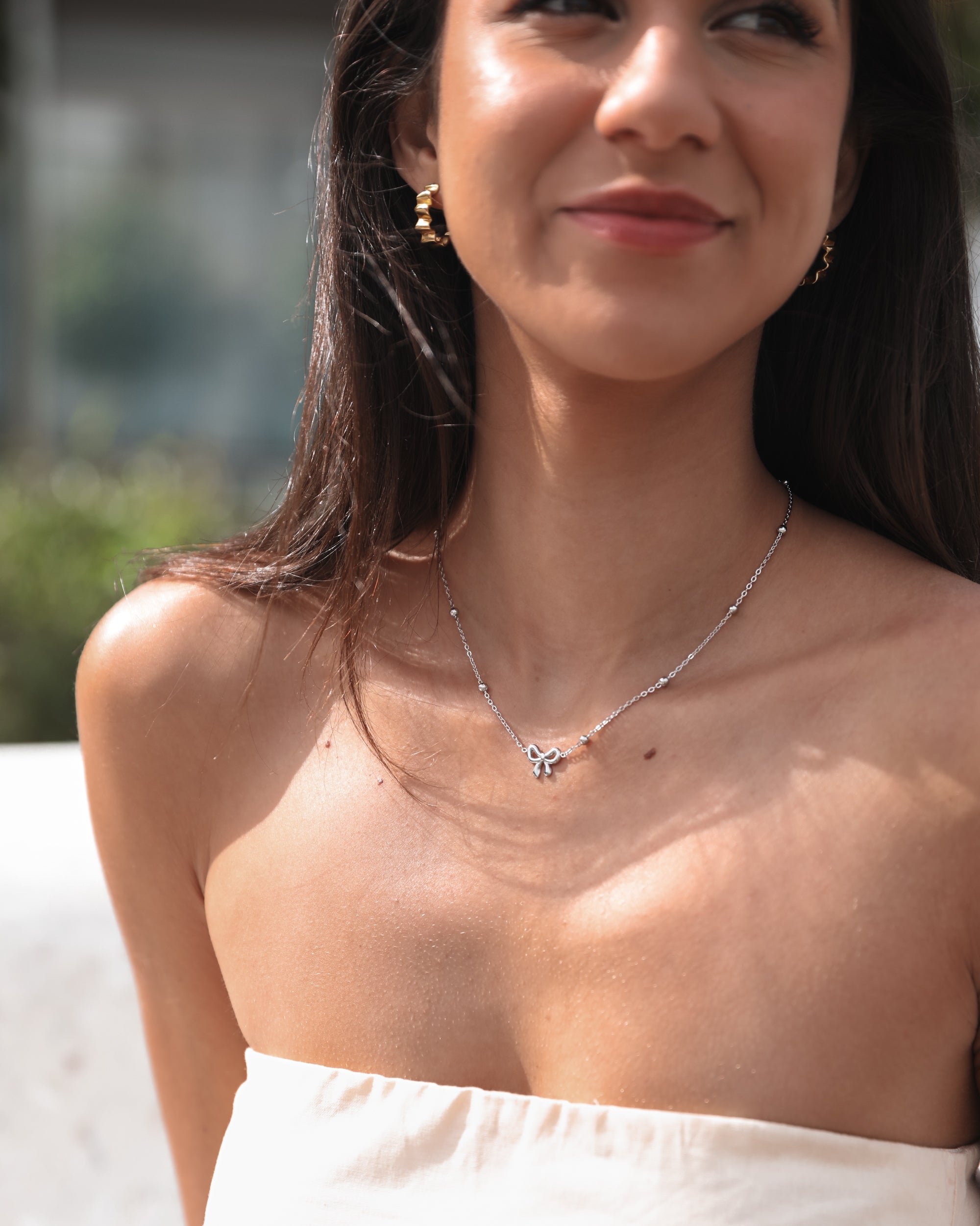 A close-up image of a woman's neckline, showcasing a silver-colored necklace with a chain and bow-shaped pendant.