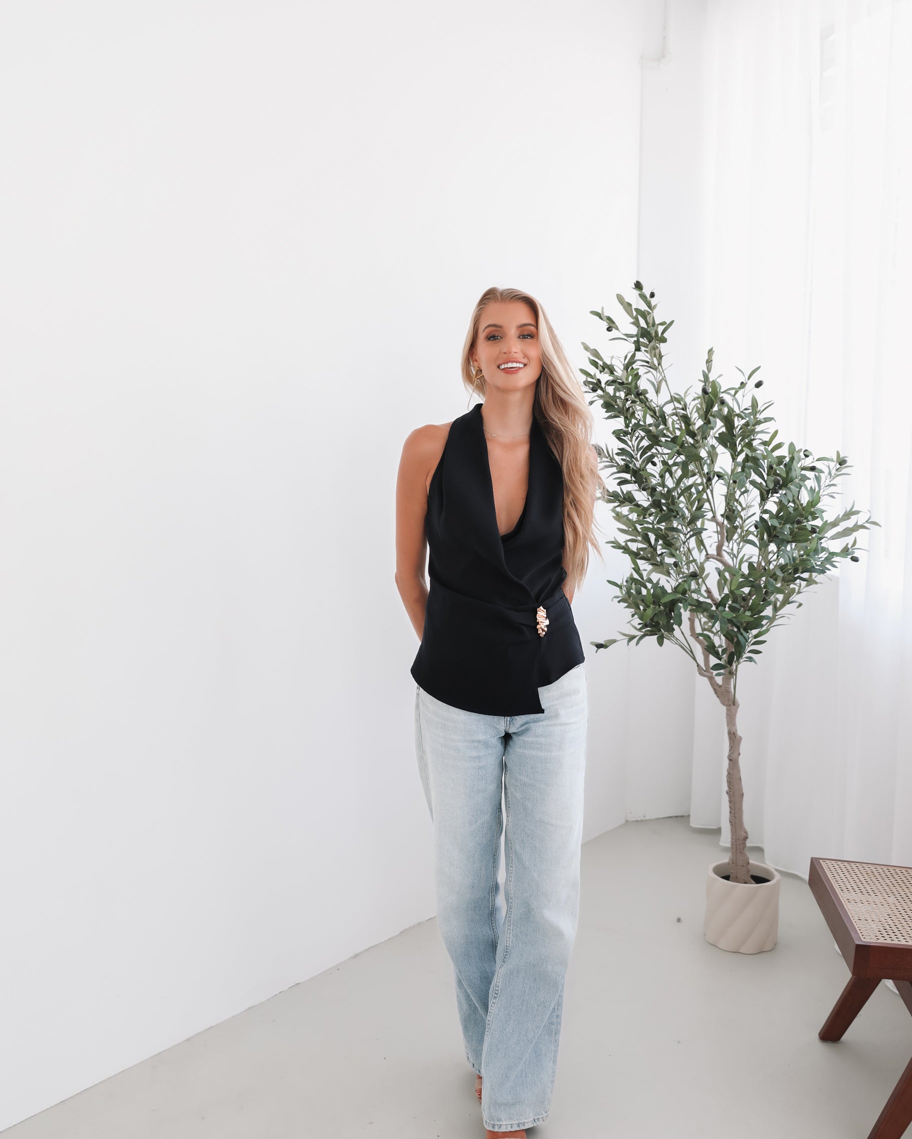 A woman wearing a navy sleeveless top with a plunge neckline and overlap bodice, paired with light jeans. She has blonde hair and is smiling.