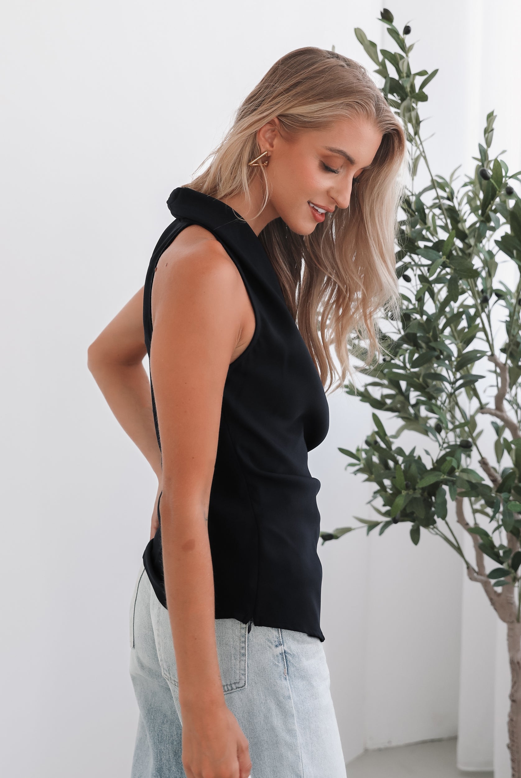 A woman wearing a navy sleeveless top with a plunge neckline and overlap bodice, paired with light jeans. She has blonde hair and is smiling.