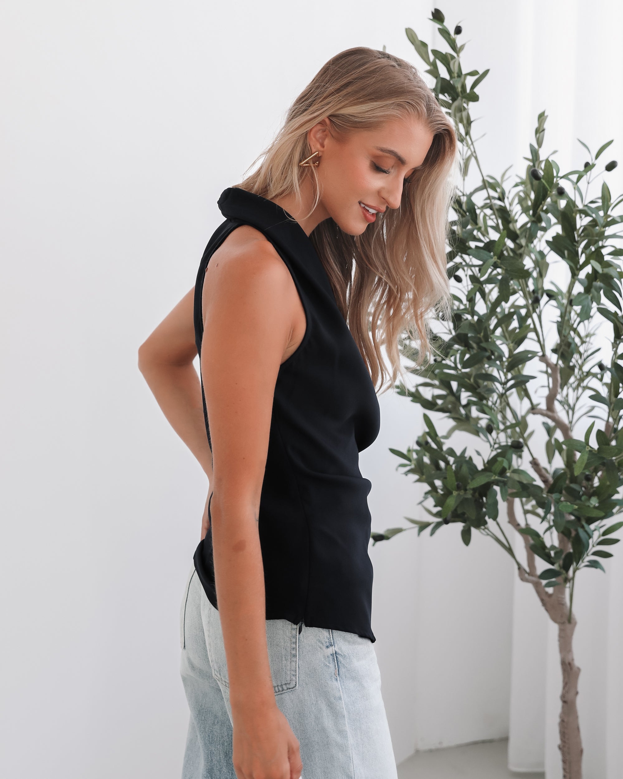 A woman wearing a navy sleeveless top with a plunge neckline and overlap bodice, paired with light jeans. She has blonde hair and is smiling.