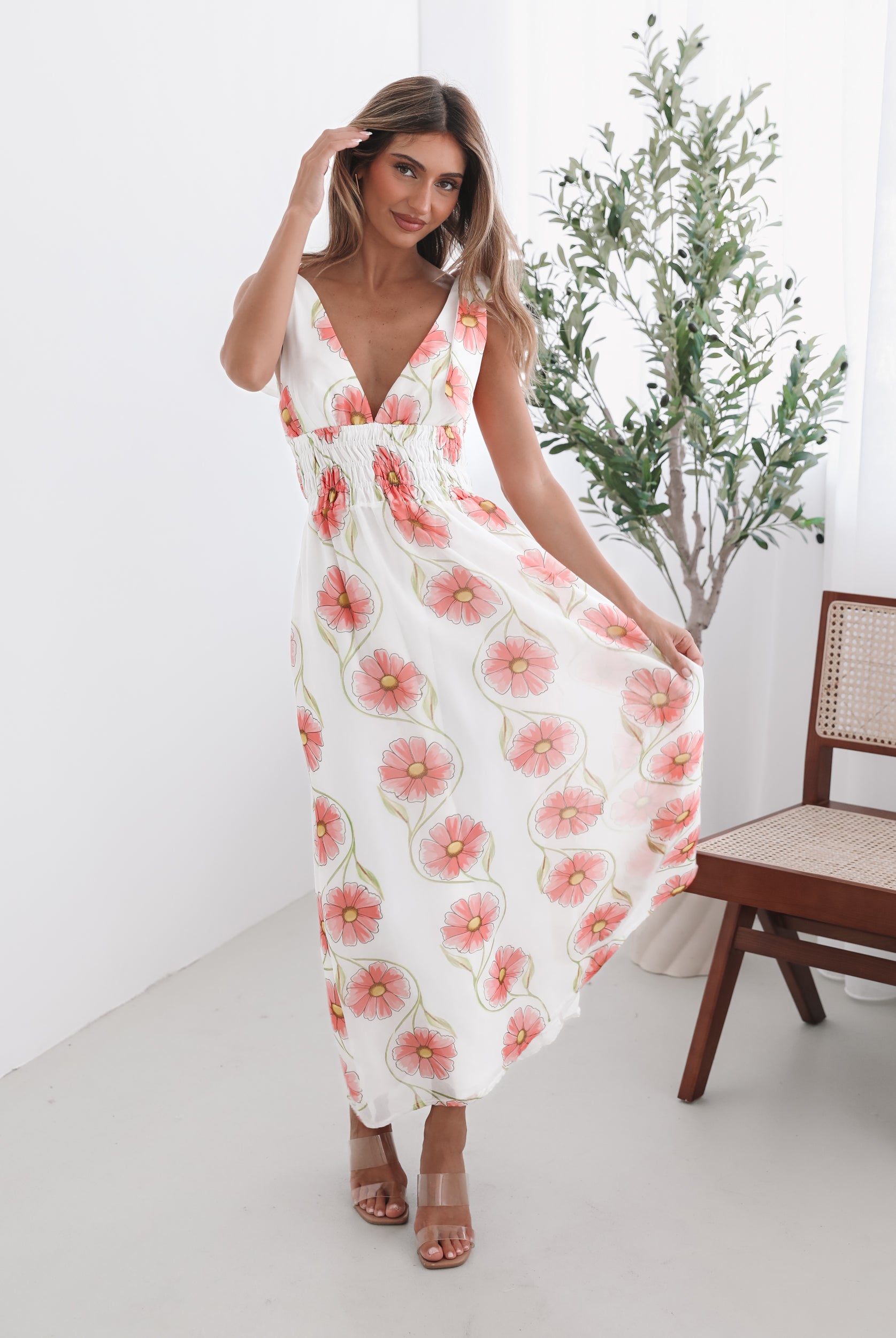 A woman standing in a room with a white floral maxi dress with bow straps and a plunge neckline.