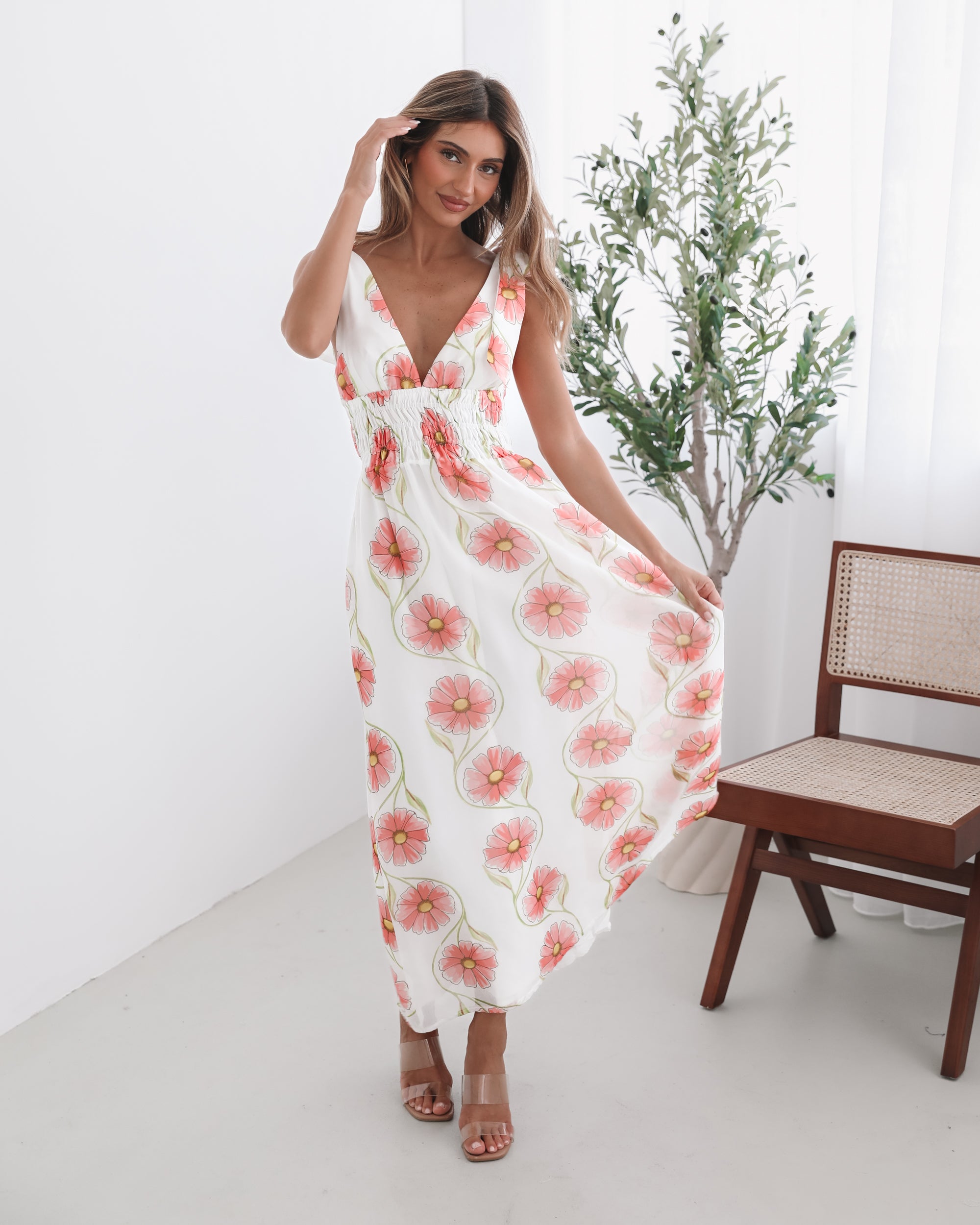 A woman standing in a room with a white floral maxi dress with bow straps and a plunge neckline.