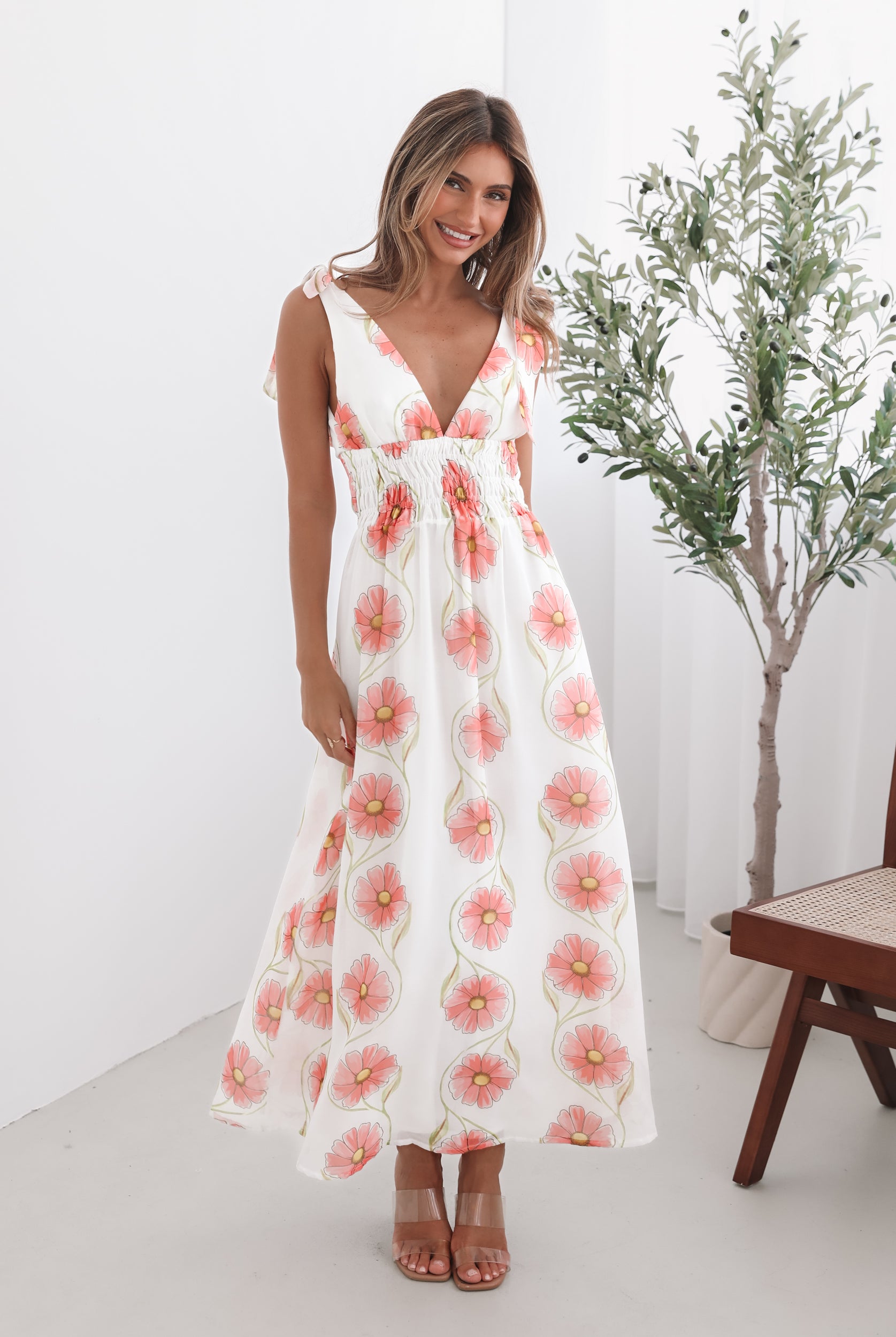 A woman standing in a room with a white floral maxi dress with bow straps and a plunge neckline.