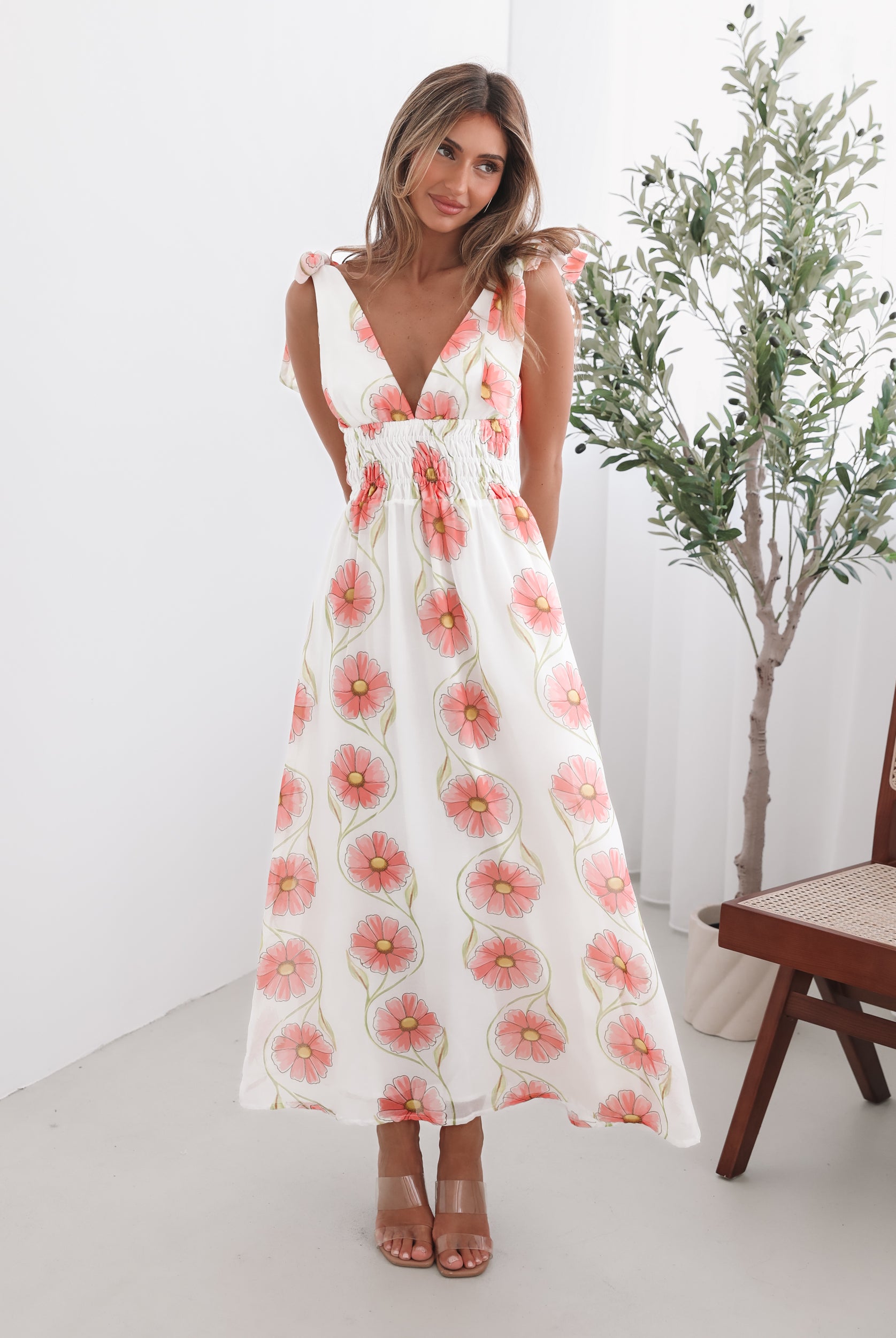 A woman standing in a room with a white floral maxi dress with bow straps and a plunge neckline.