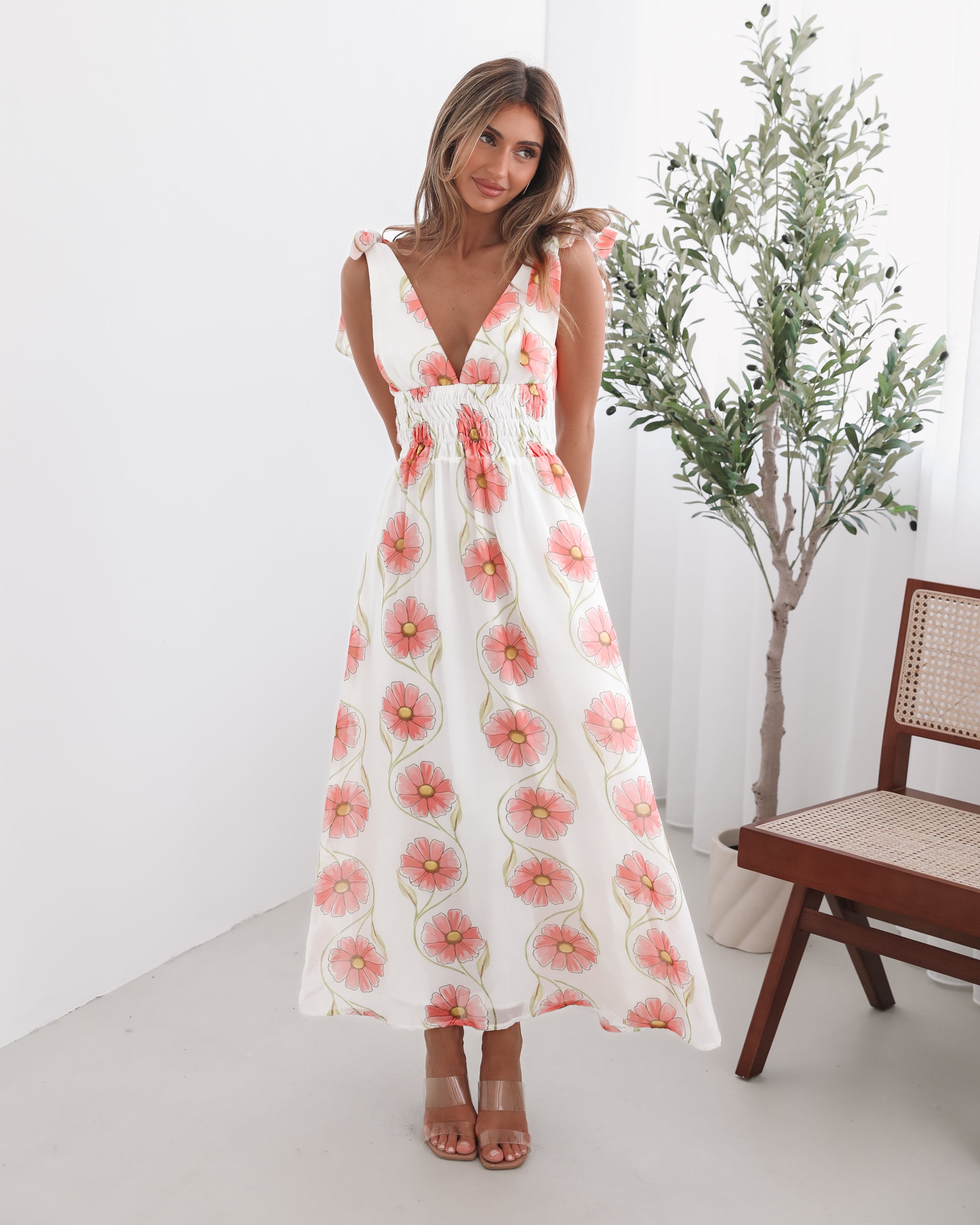 A woman standing in a room with a white floral maxi dress with bow straps and a plunge neckline.