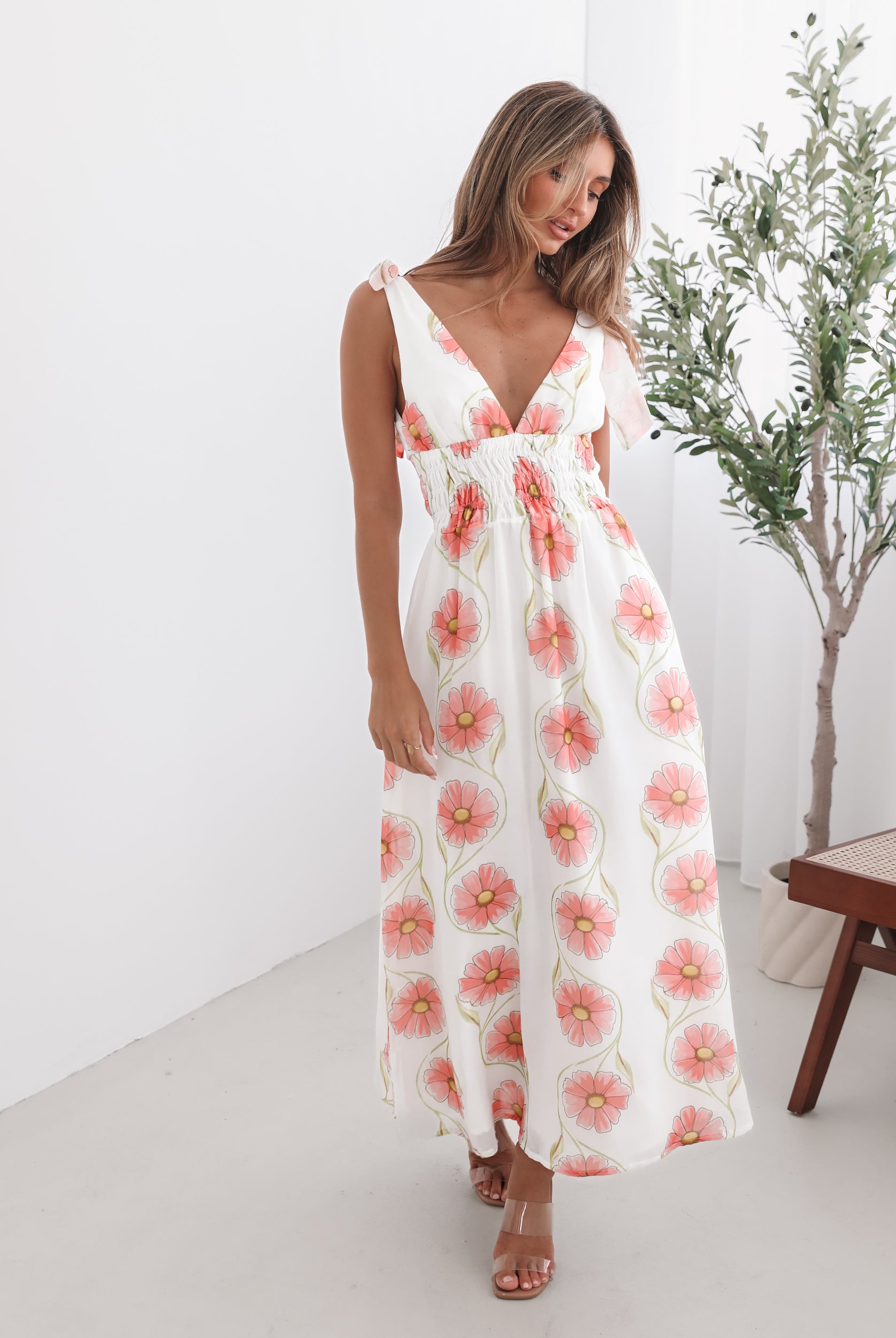 A woman standing in a room with a white floral maxi dress with bow straps and a plunge neckline.