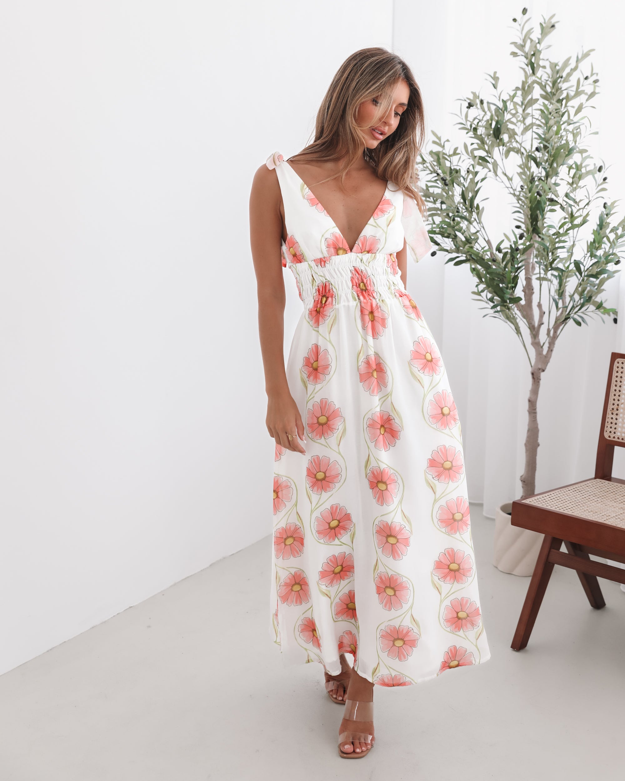 A woman standing in a room with a white floral maxi dress with bow straps and a plunge neckline.