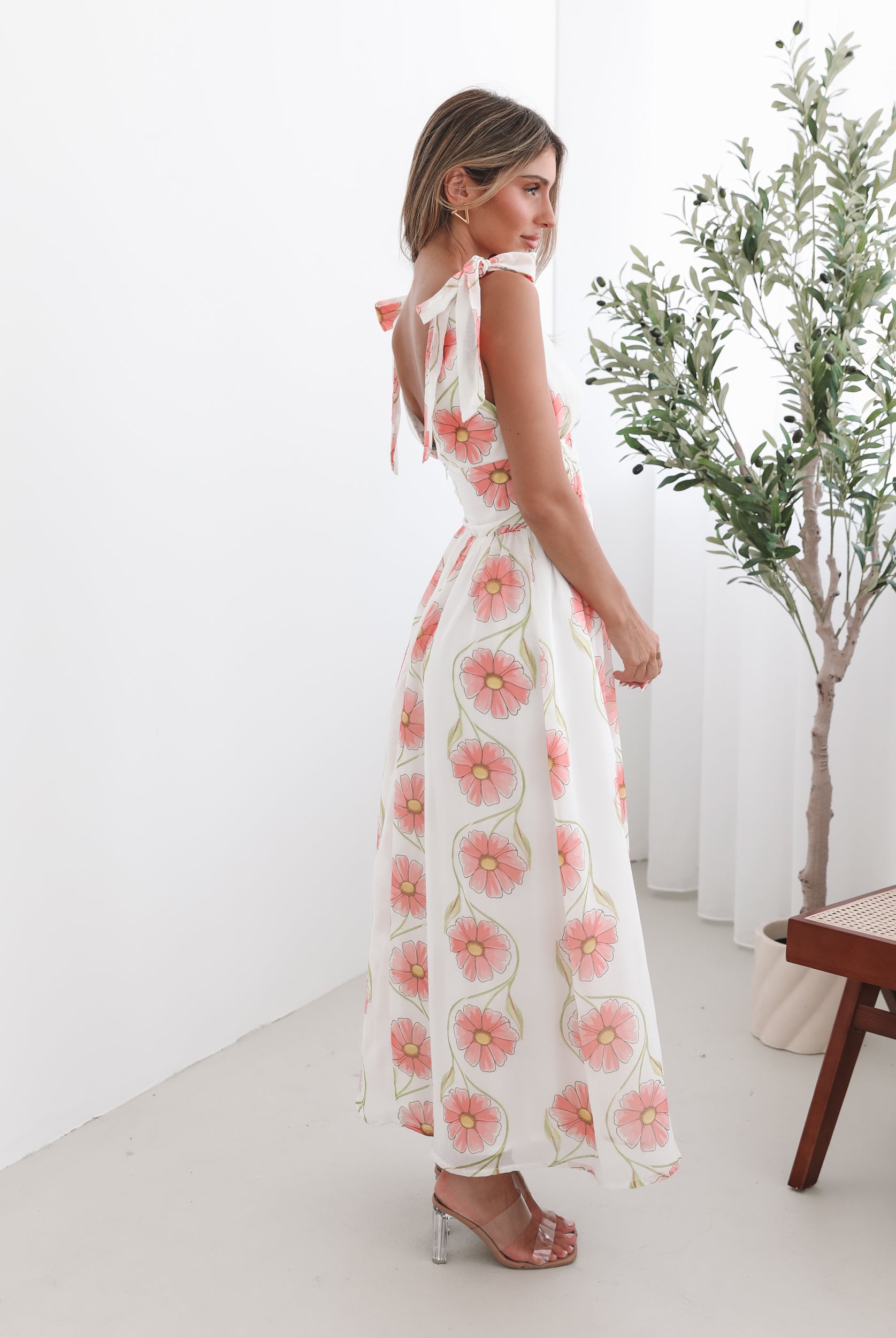 A woman standing in a room with a white floral maxi dress with bow straps and a plunge neckline.
