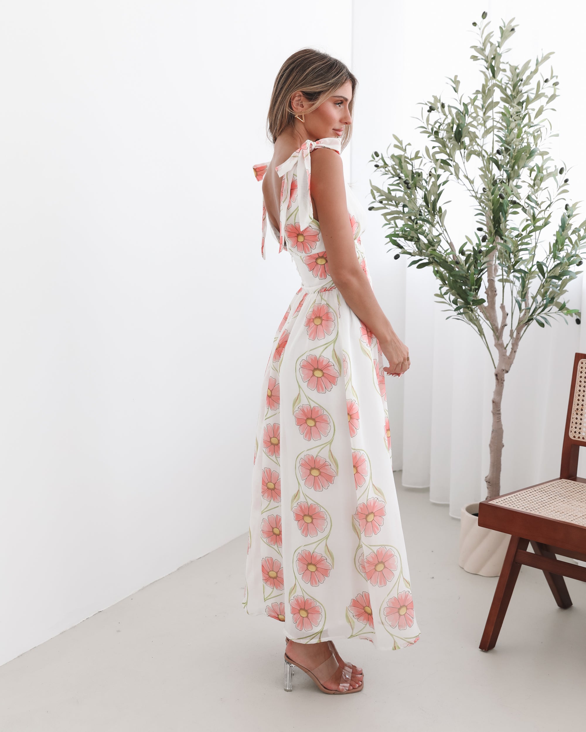 A woman standing in a room with a white floral maxi dress with bow straps and a plunge neckline.