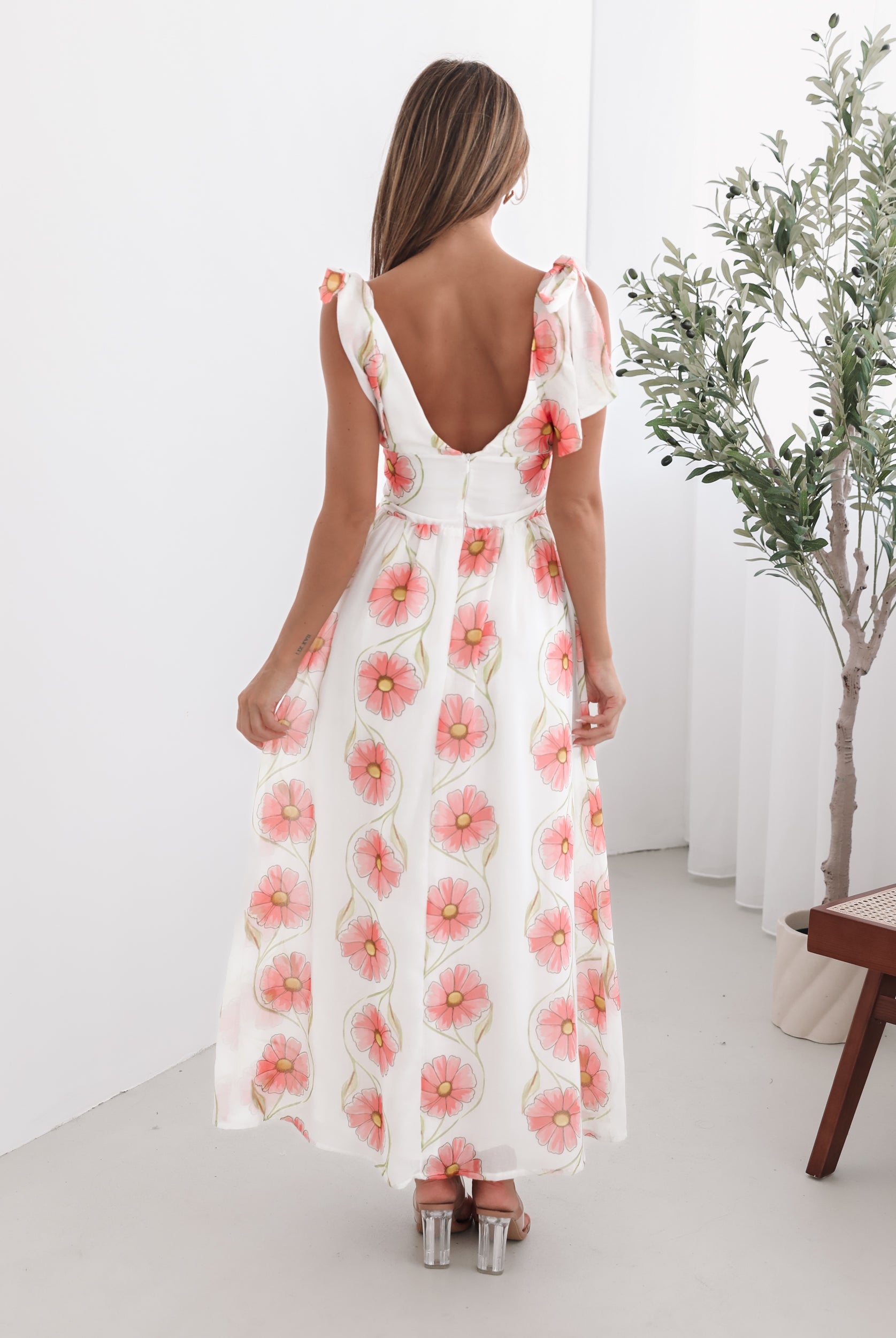 A woman standing in a room with a white floral maxi dress with bow straps and a plunge neckline.